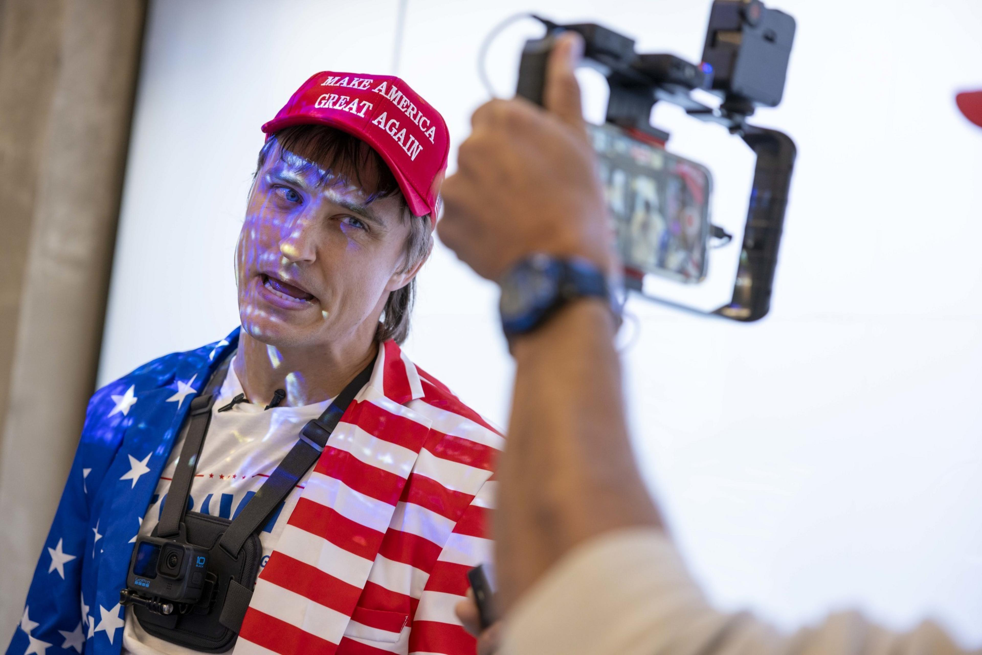A person wearing a red "Make America Great Again" cap and an American flag-patterned jacket is being filmed with a smartphone mounted on a stabilizer.