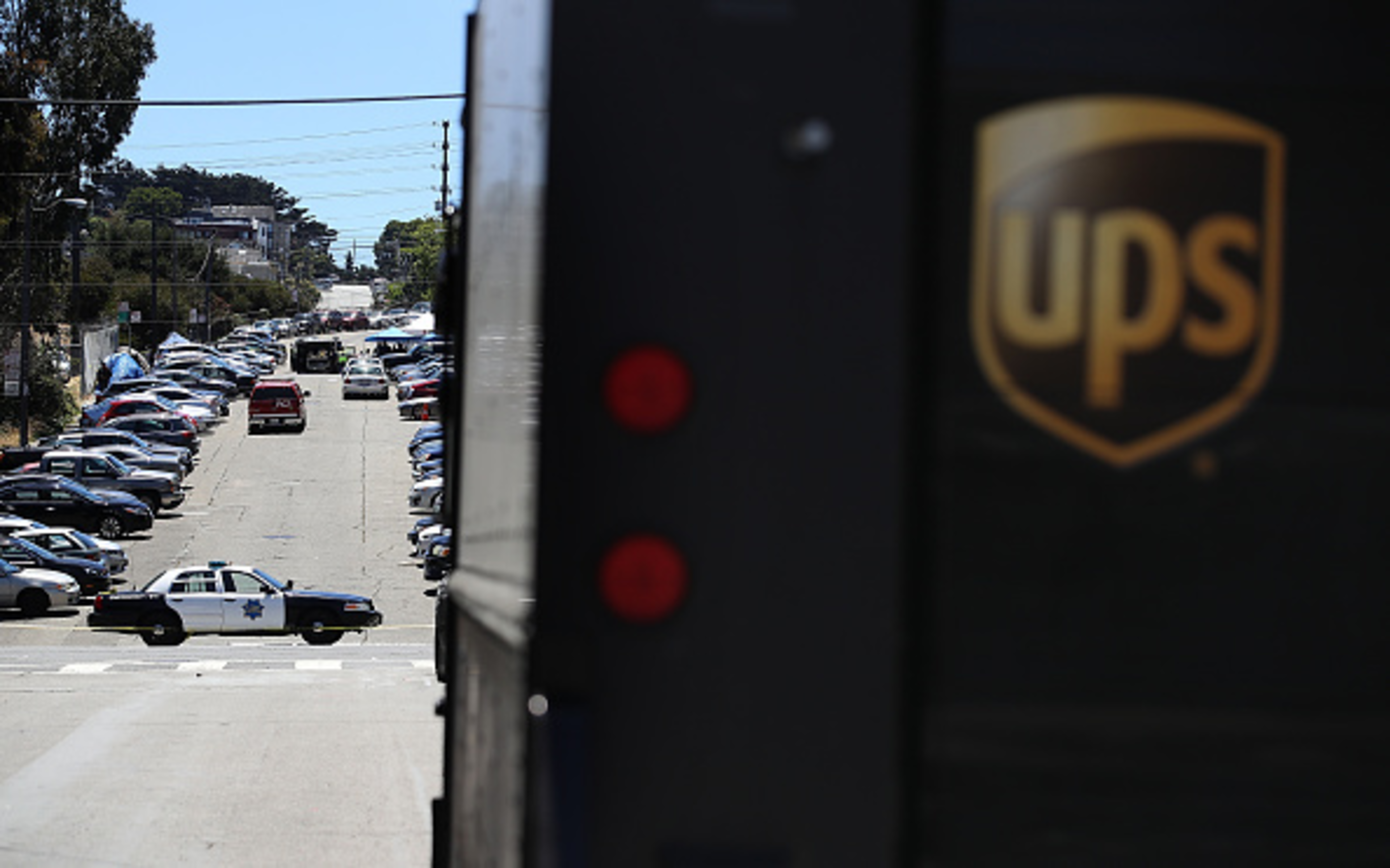 A police car blocks a street lined with parked cars, partially obscured by a UPS truck in the foreground on the right.