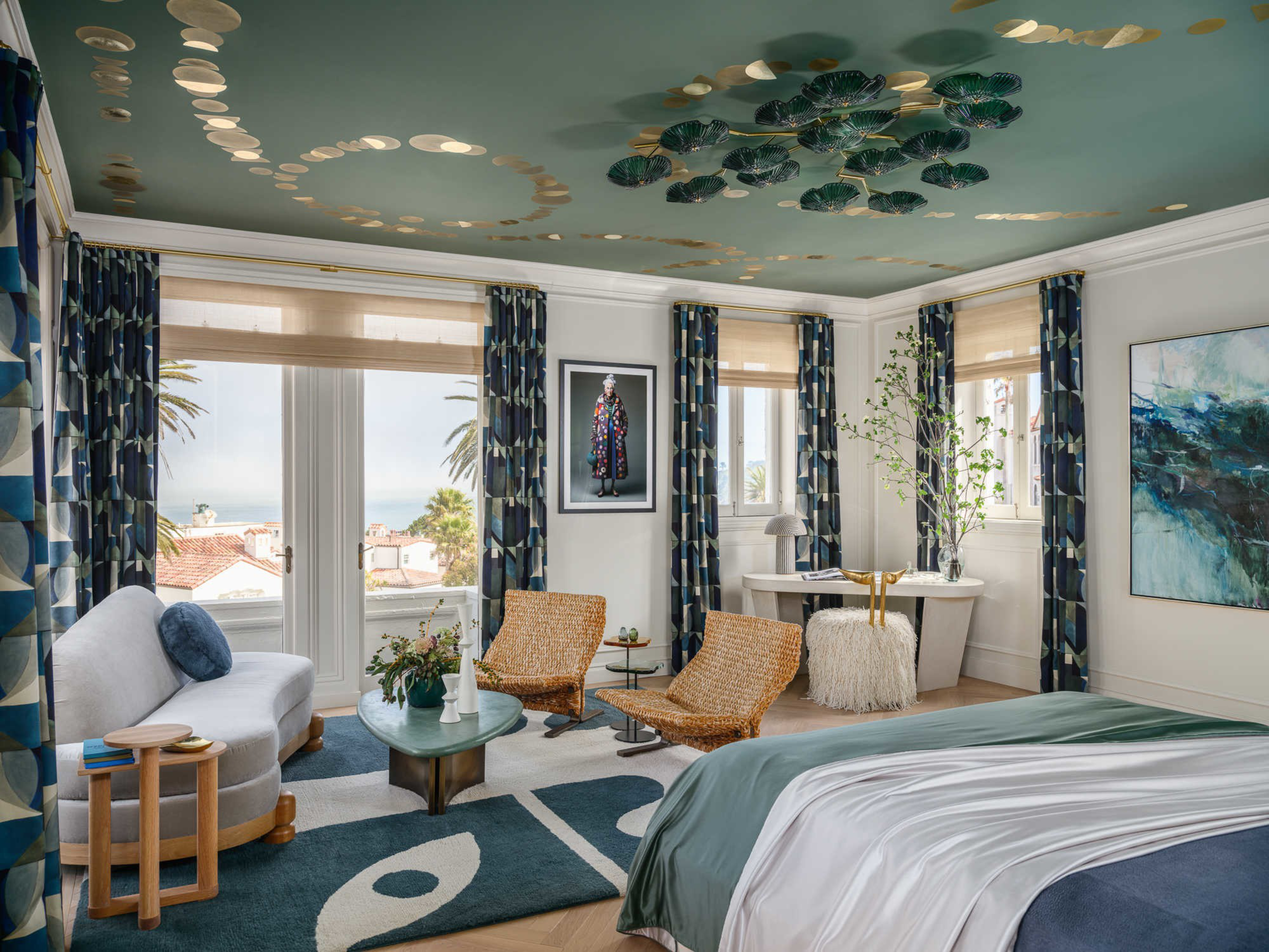 A spacious bedroom features a green ceiling with gold and leaf designs, a bed with green and white bedding, wicker chairs, blue-patterned curtains, and large windows with a sea view.