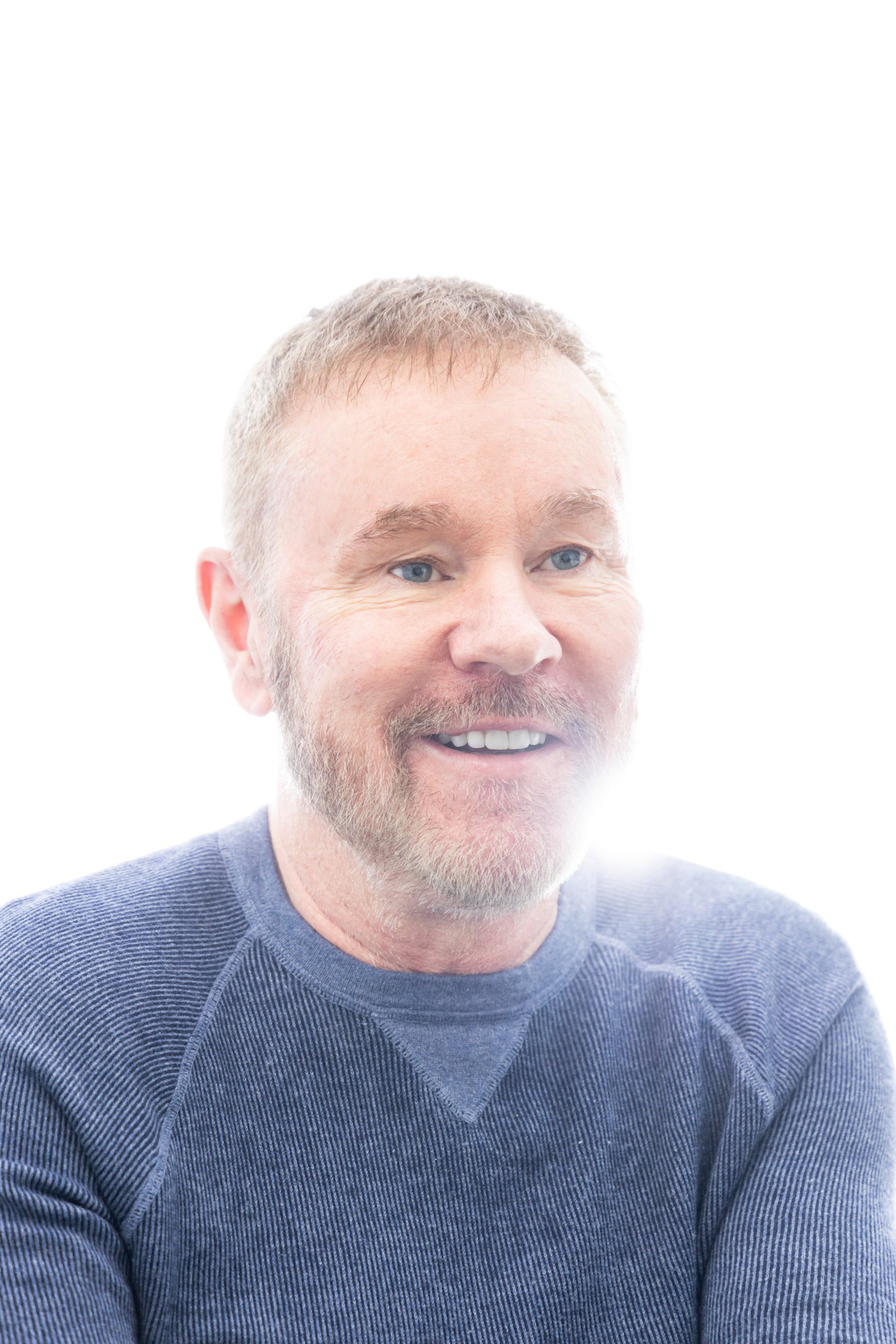 A person with a short beard is smiling, wearing a textured blue sweater. The image is bright with a high-key background, illuminating their face.
