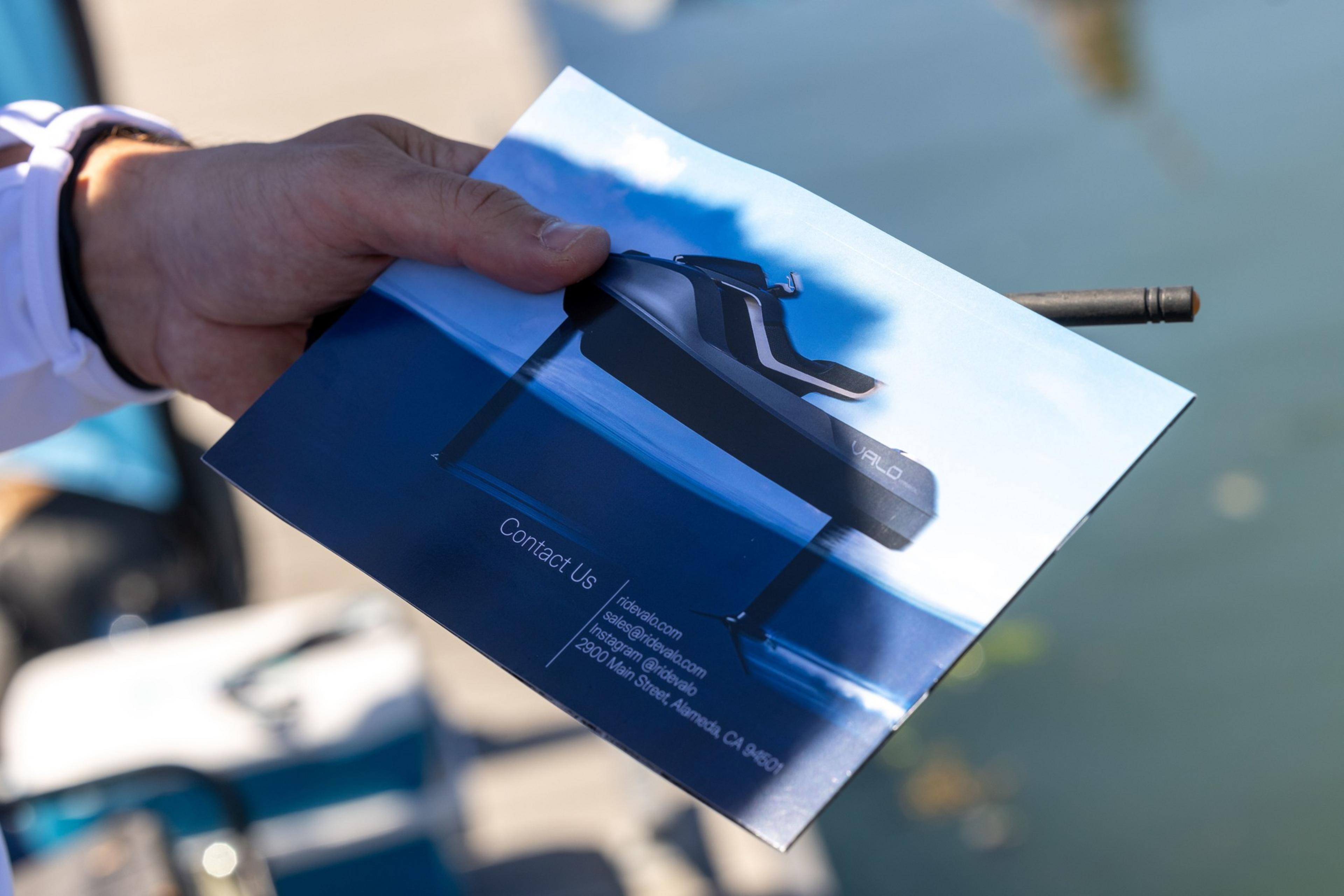 A person is holding a brochure featuring a black watercraft called &quot;VALO,&quot; floating above water. Contact details, including a website, email, Instagram, and address, are shown below.