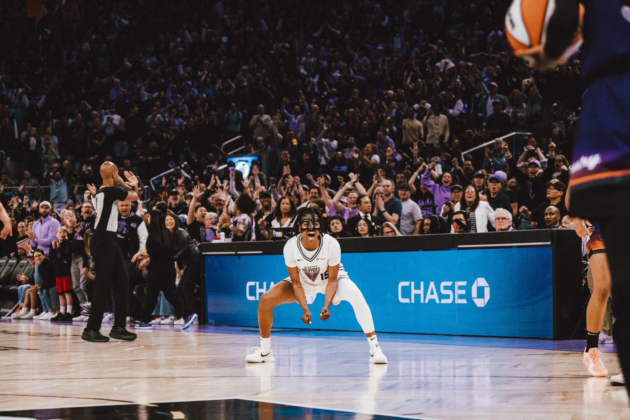 A basketball player in white celebrates intensely on the court while a large crowd cheers in the background during a game.