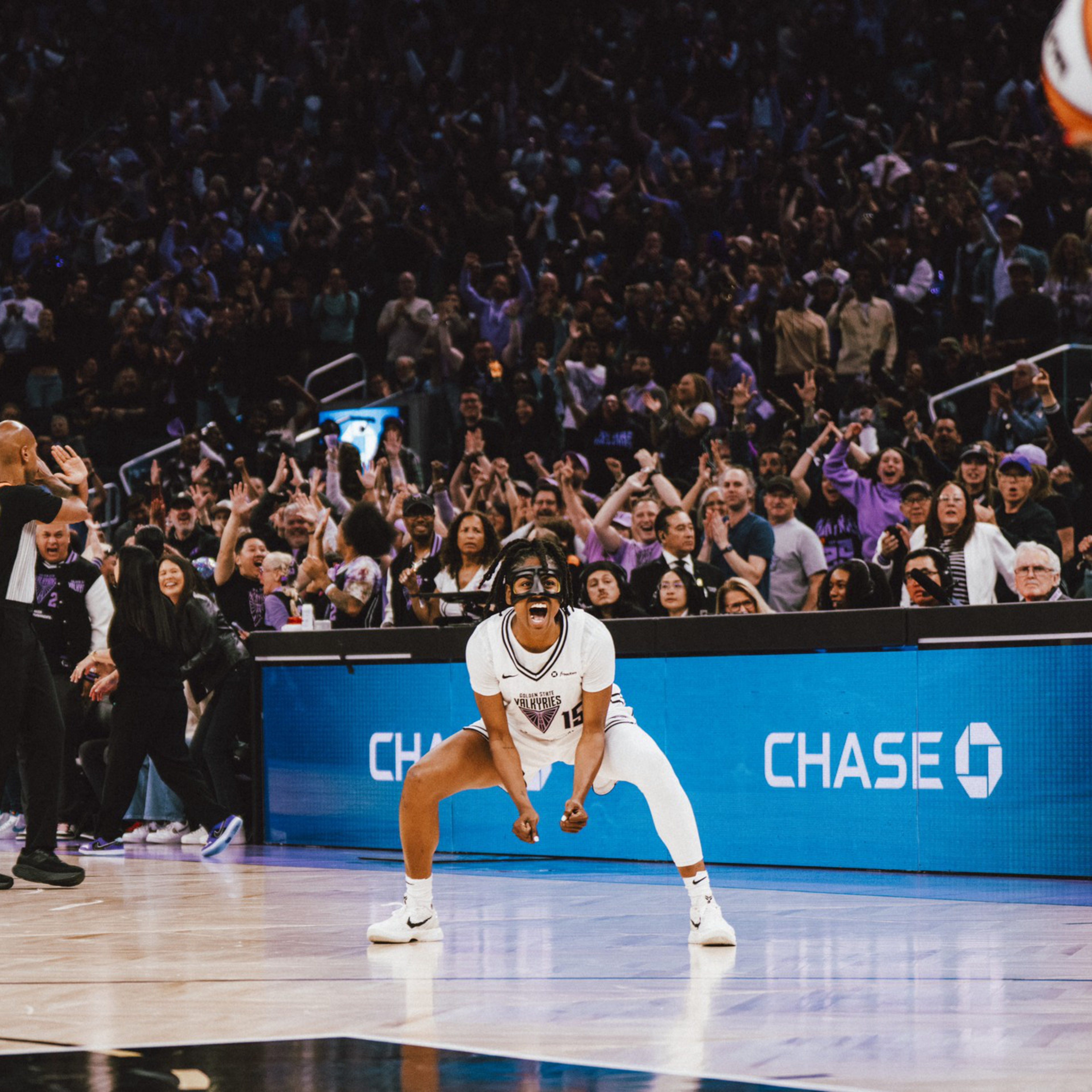 A basketball player in white celebrates intensely on the court while a large crowd cheers in the background during a game.