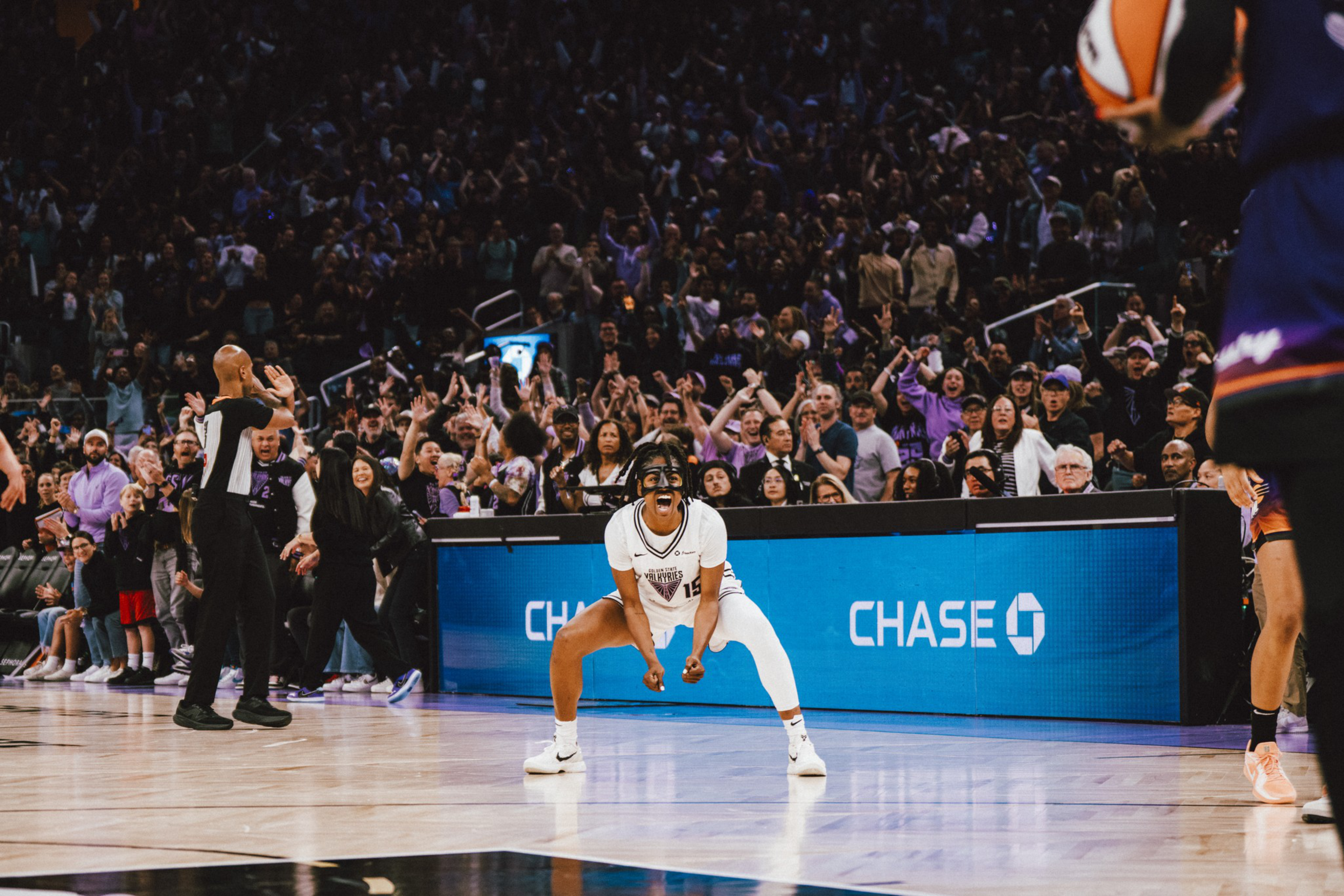 A basketball player in white celebrates intensely on the court while a large crowd cheers in the background during a game.