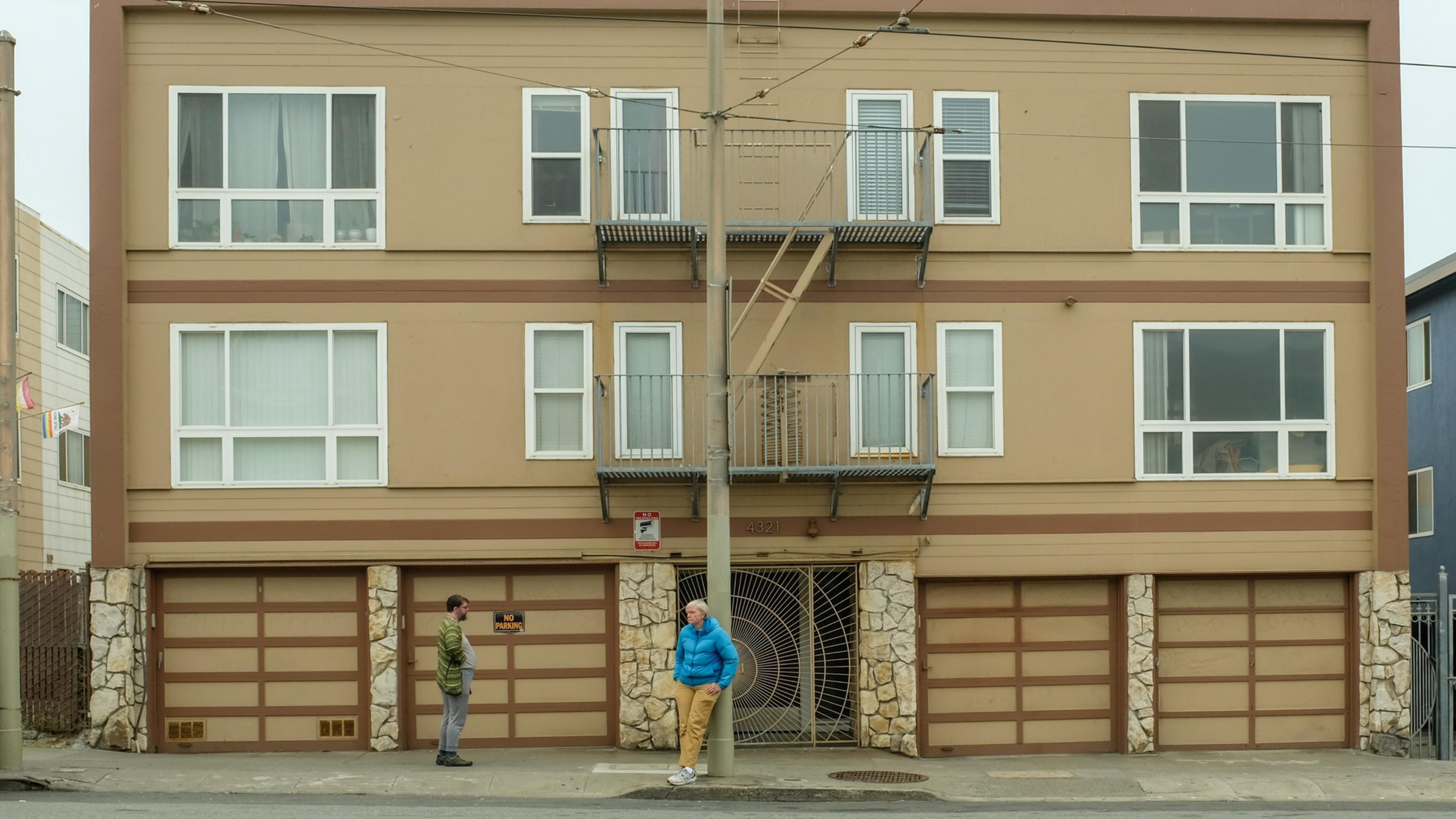 Two people stand on the sidewalk in front of a brown, three-story apartment building with four garage doors and an iron gate entrance.
