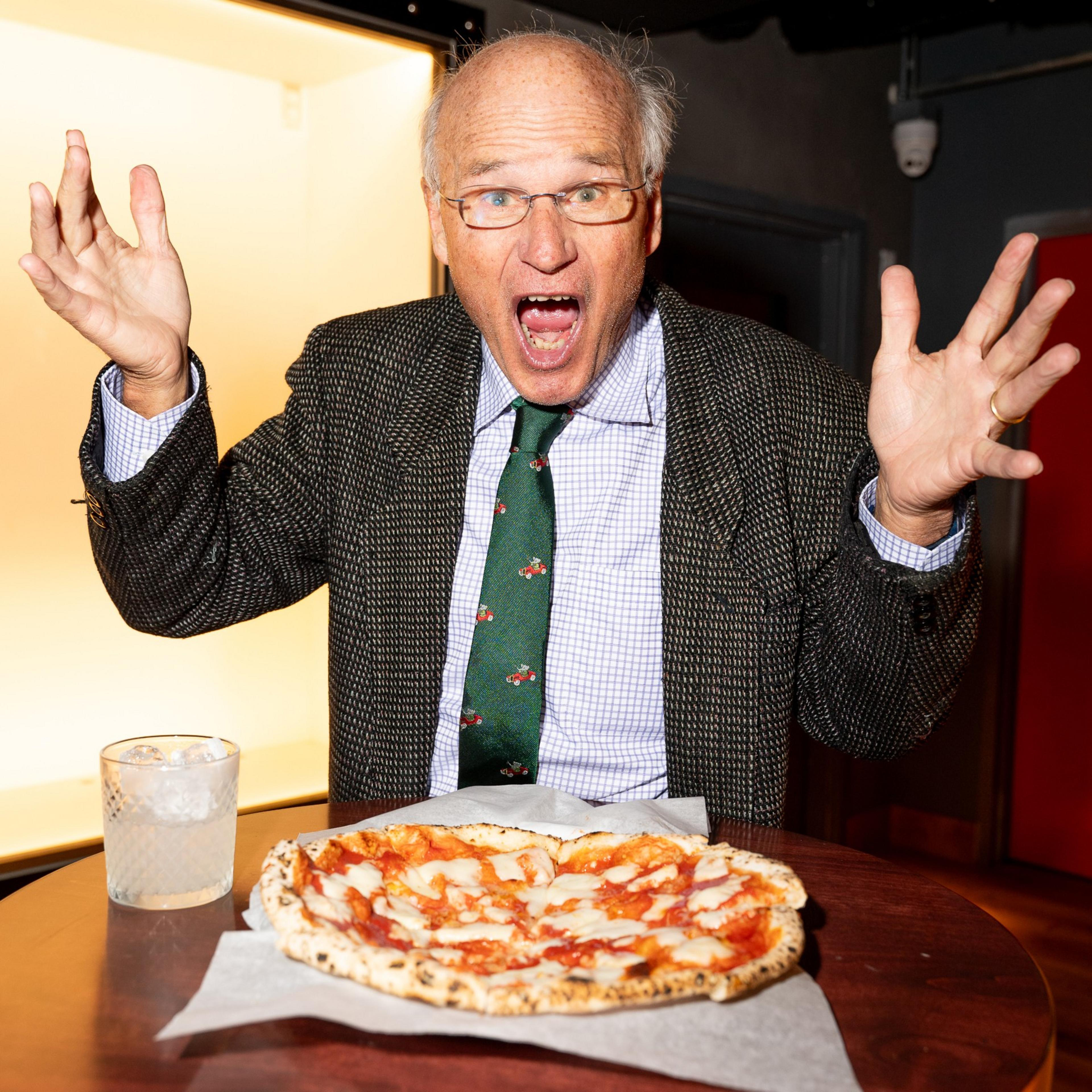 A man in a suit and tie, expressing surprise or excitement, is seated at a table with a pizza and a glass of ice water in front of him.