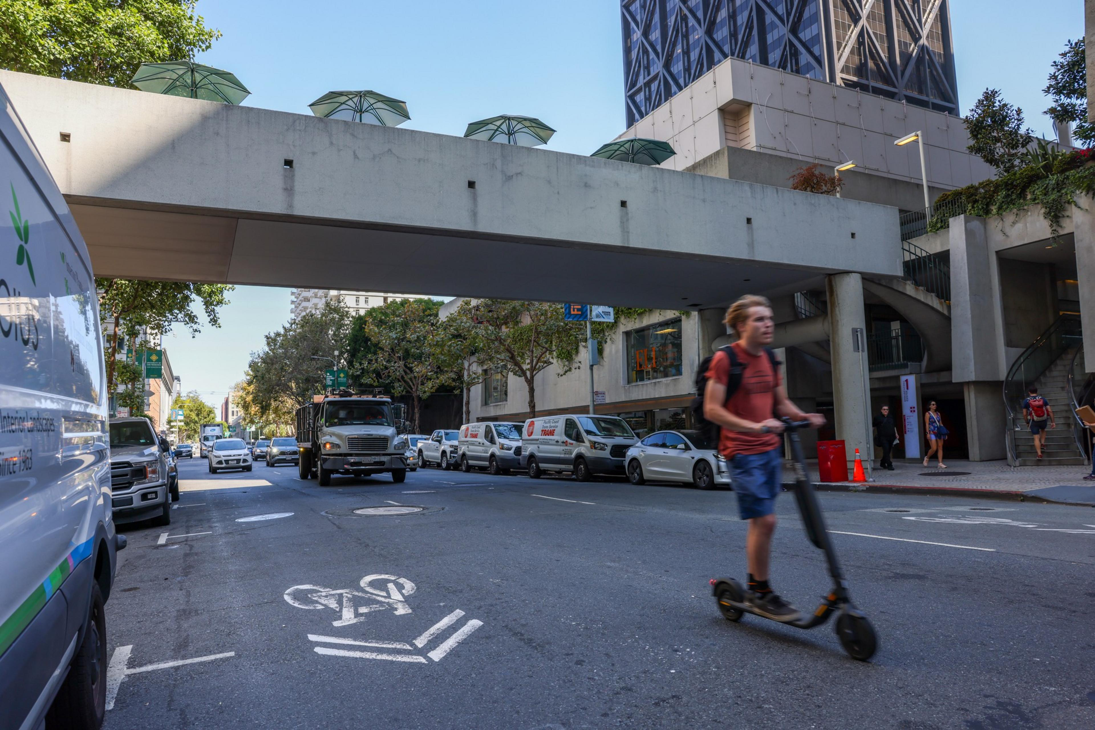 A person rides an electric scooter on a city street with cars parked alongside and a large white overpass above.