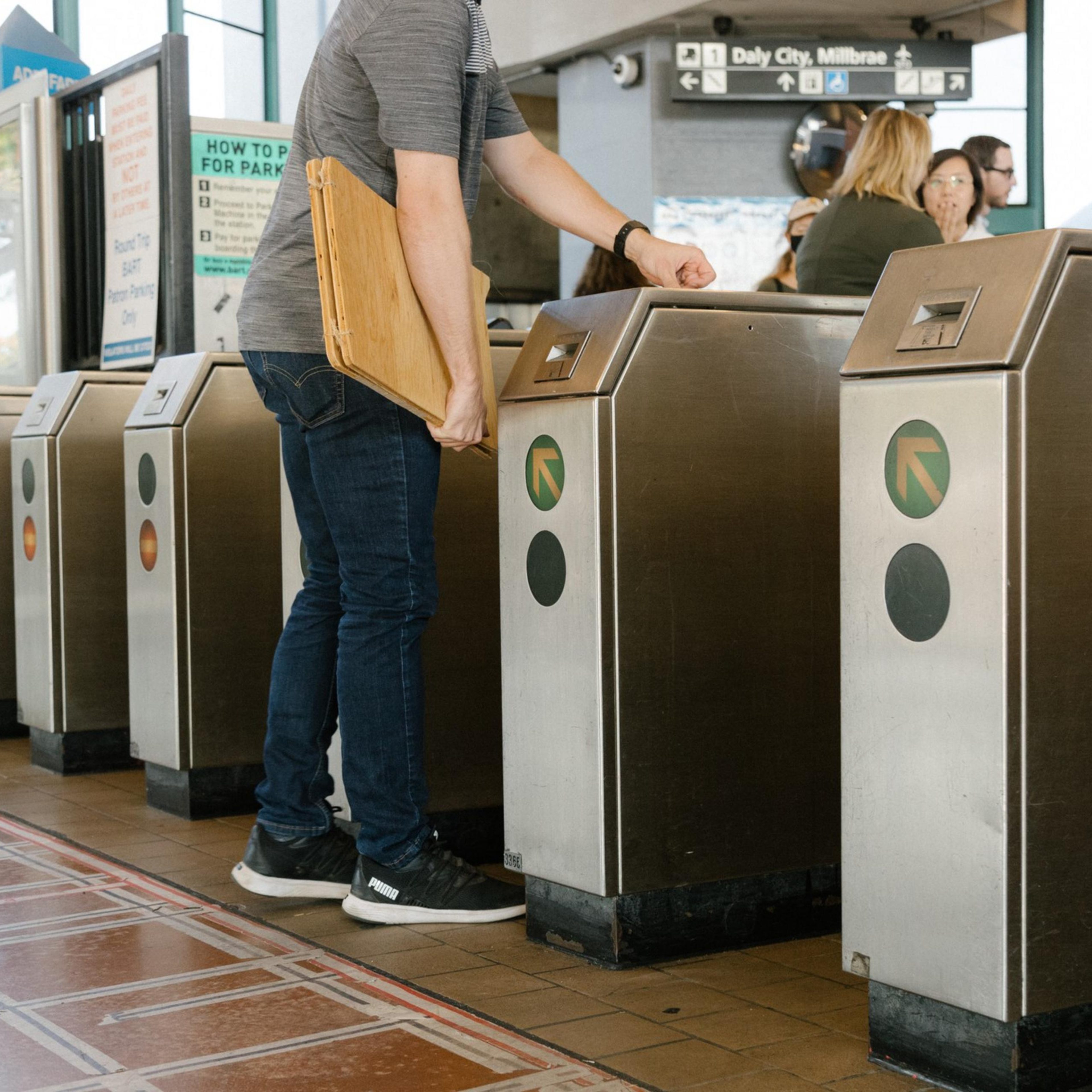 A person scans a card at a turnstile in a transit station, with other turnstiles and a sign showing directions to Daly City and Millbrae in the background.