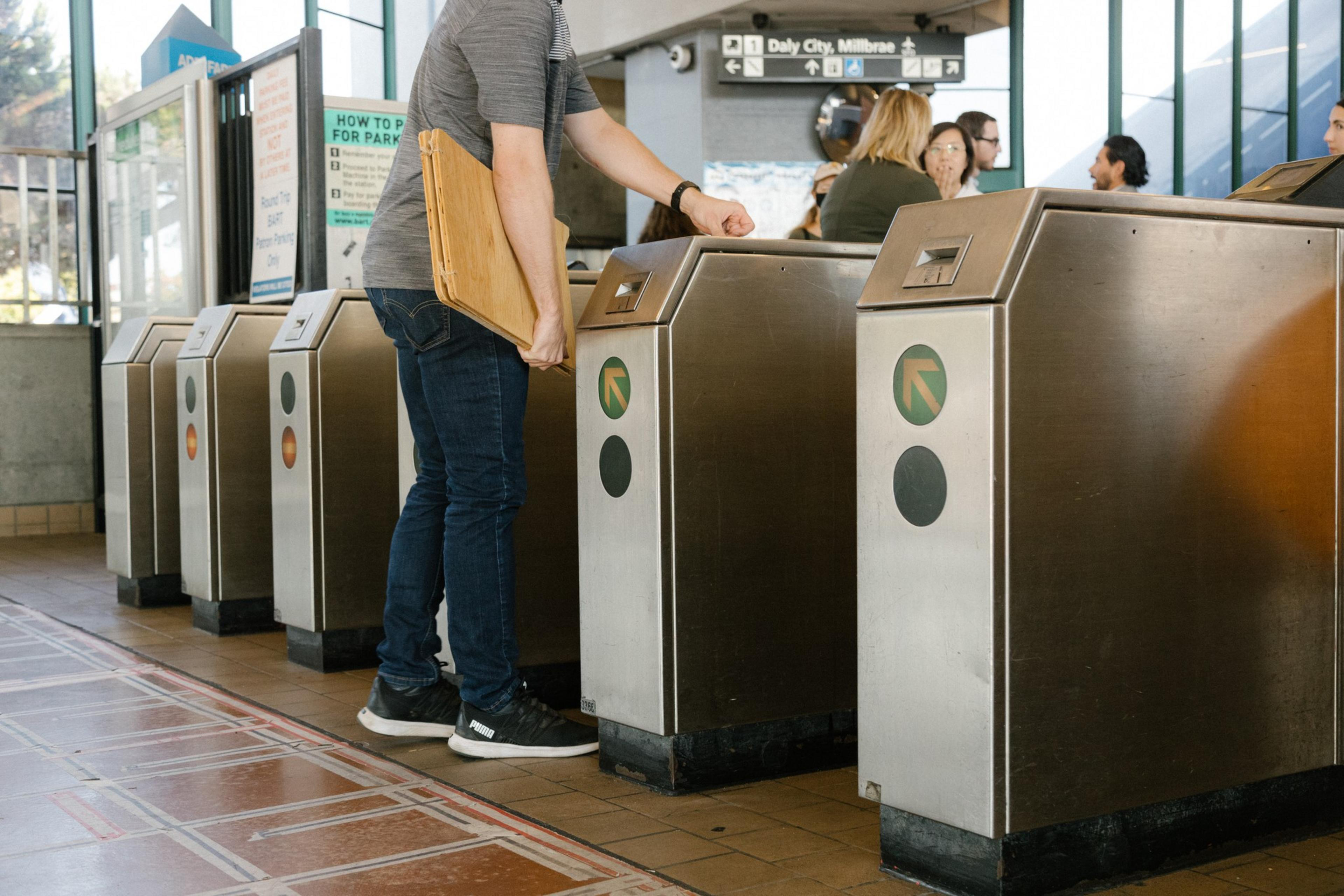 A person scans a card at a turnstile in a transit station, with other turnstiles and a sign showing directions to Daly City and Millbrae in the background.