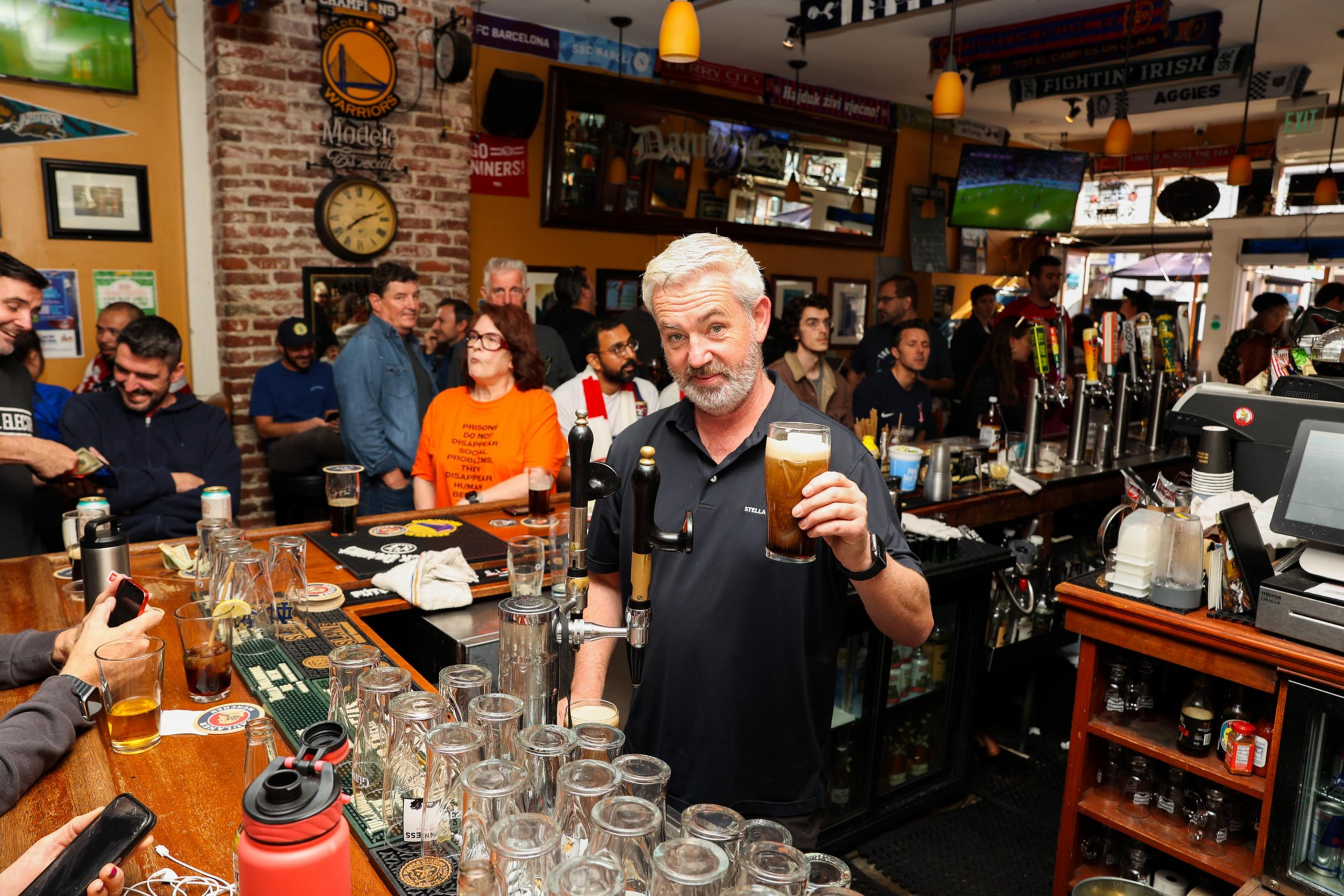 A man with gray hair behind a bar holds up a pint of beer, surrounded by empty glasses and a crowd of people socializing and watching sports on TVs.