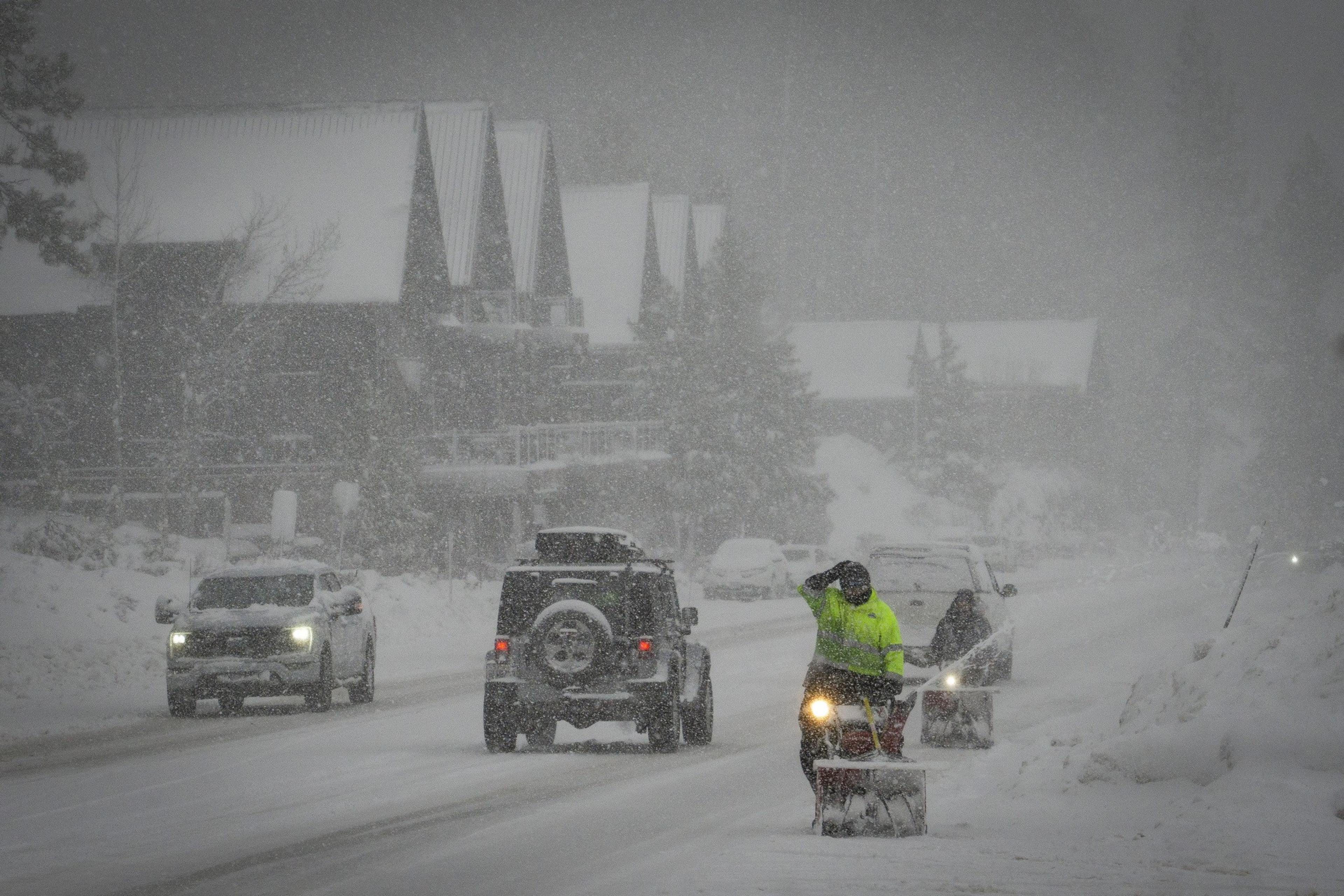 Cars drive along snowy road as workers move alongside with snow blowers
