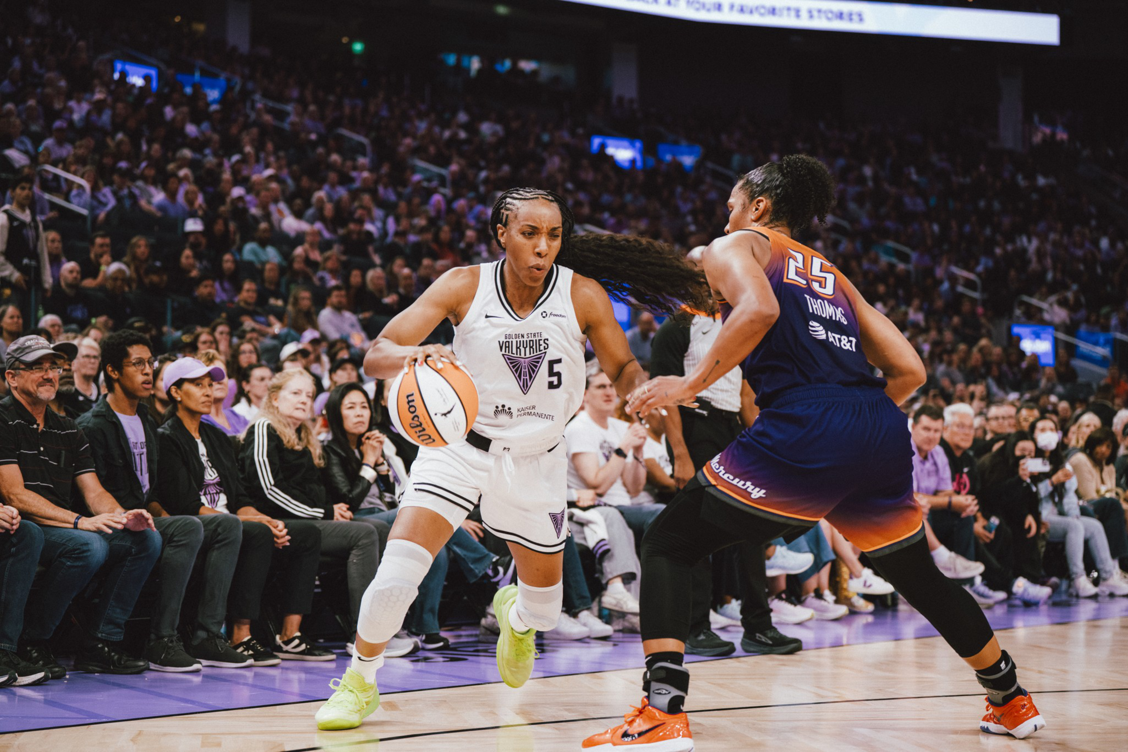 A basketball player in white dribbles past a defender in navy and orange, with a crowd watching intently in the background.