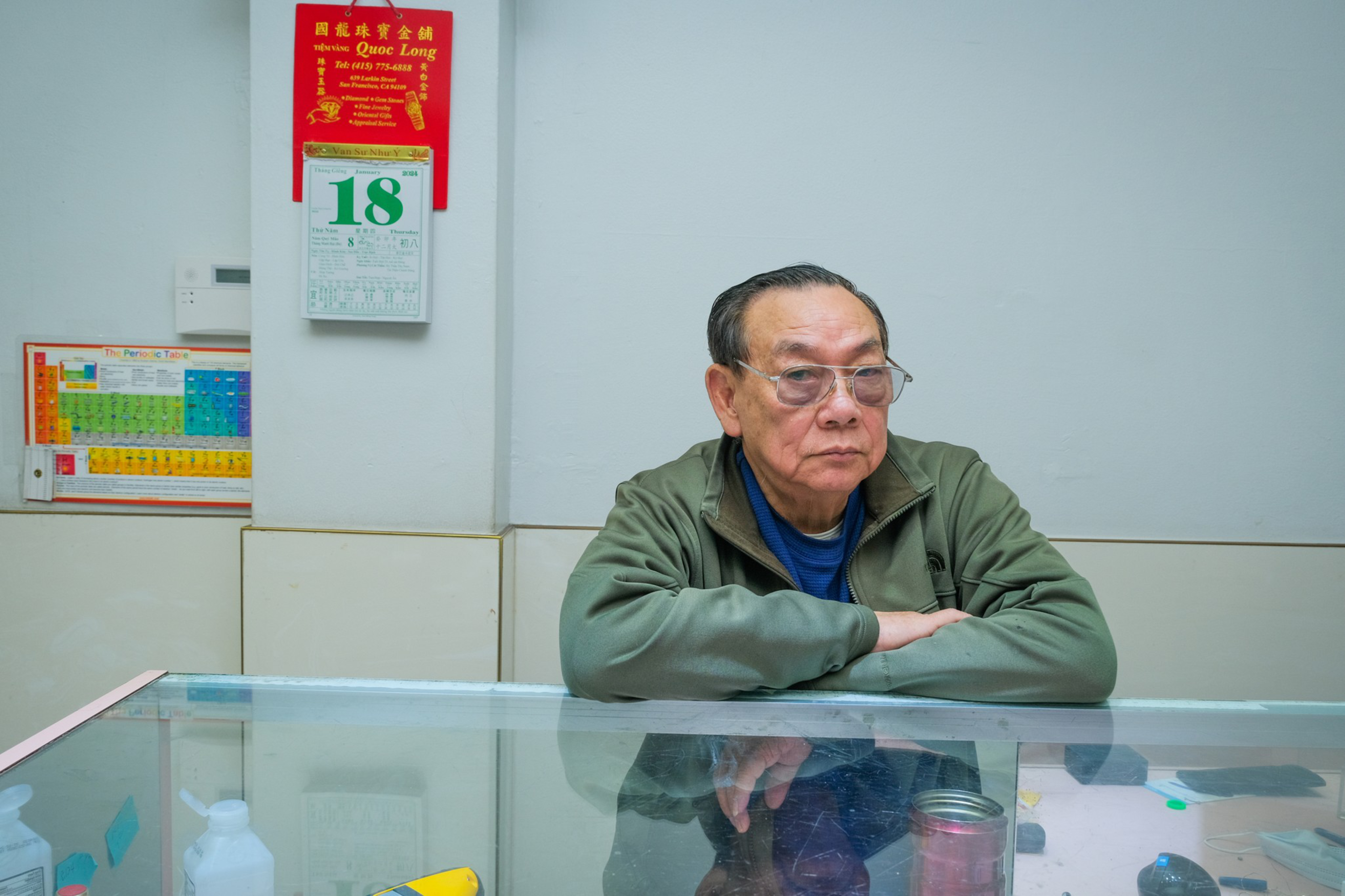 An older man with glasses sits behind a glass counter, with calendars and a chart on the wall behind him.
