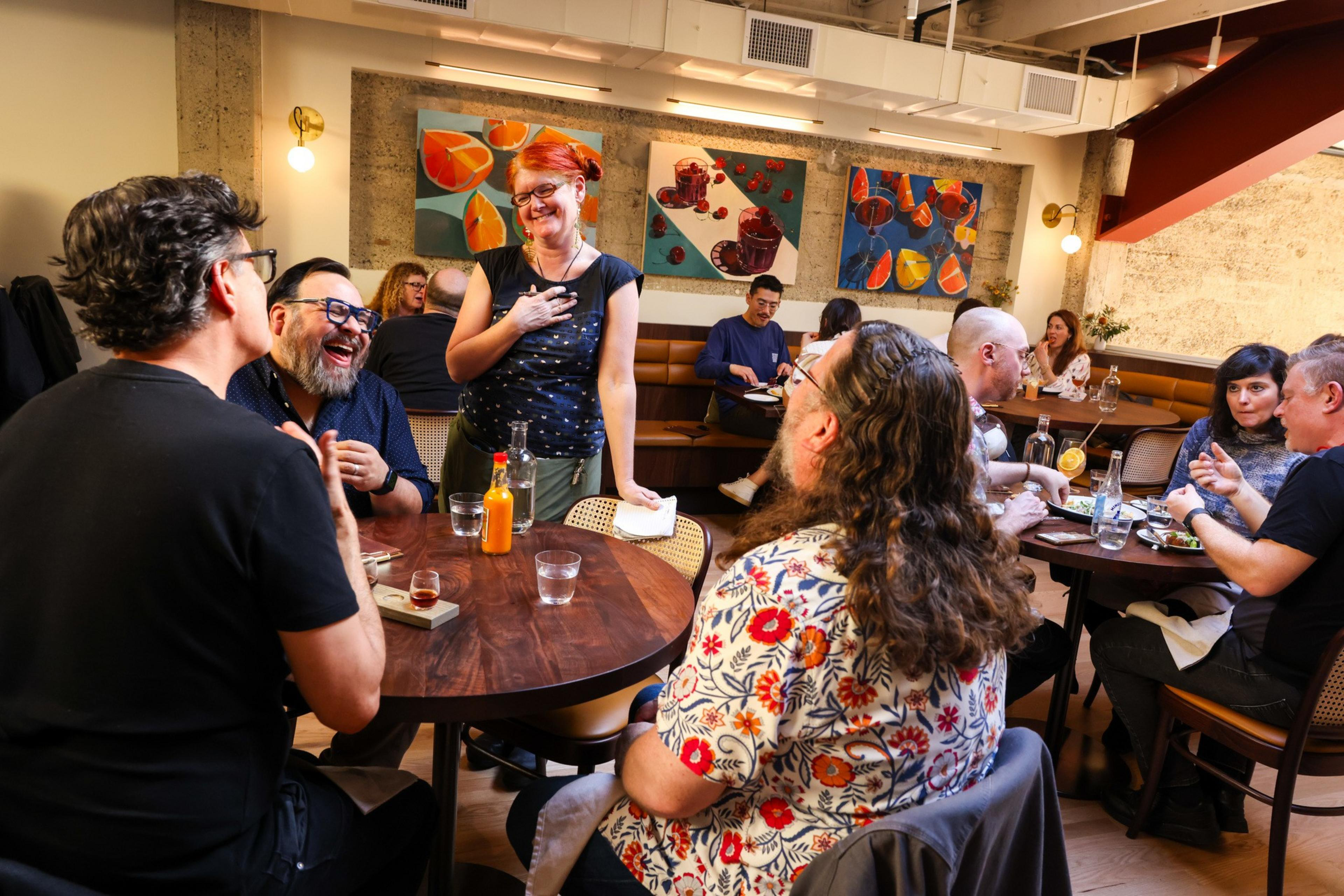 A group of people happily converse at a cozy restaurant table. The walls feature vibrant, abstract paintings. The atmosphere looks friendly and relaxed.