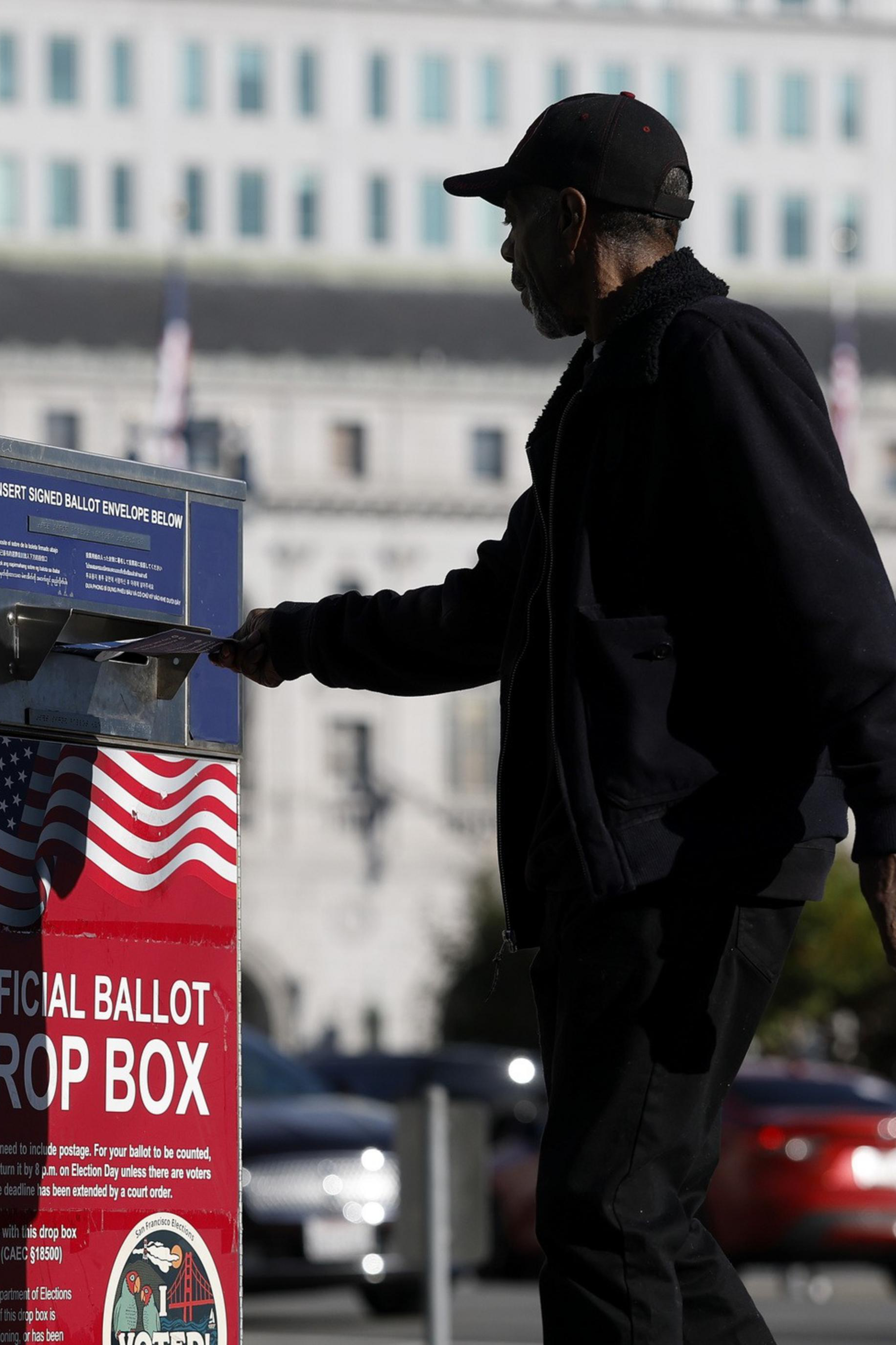 A man drops a ballot into a red, white, and blue official ballot drop box with U.S. flags, located outdoors near a large building.