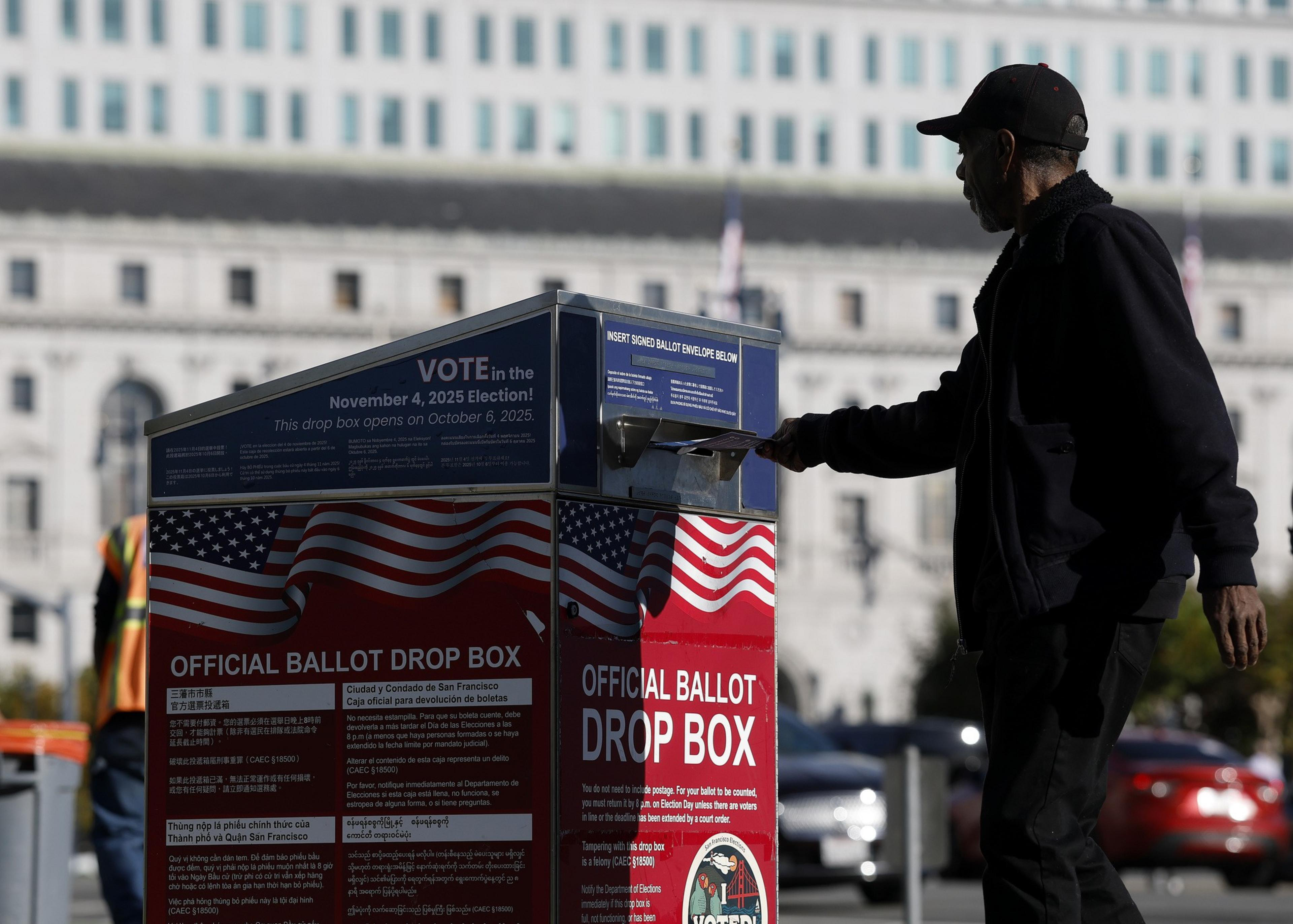 A man drops a ballot into a red, white, and blue official ballot drop box with U.S. flags, located outdoors near a large building.