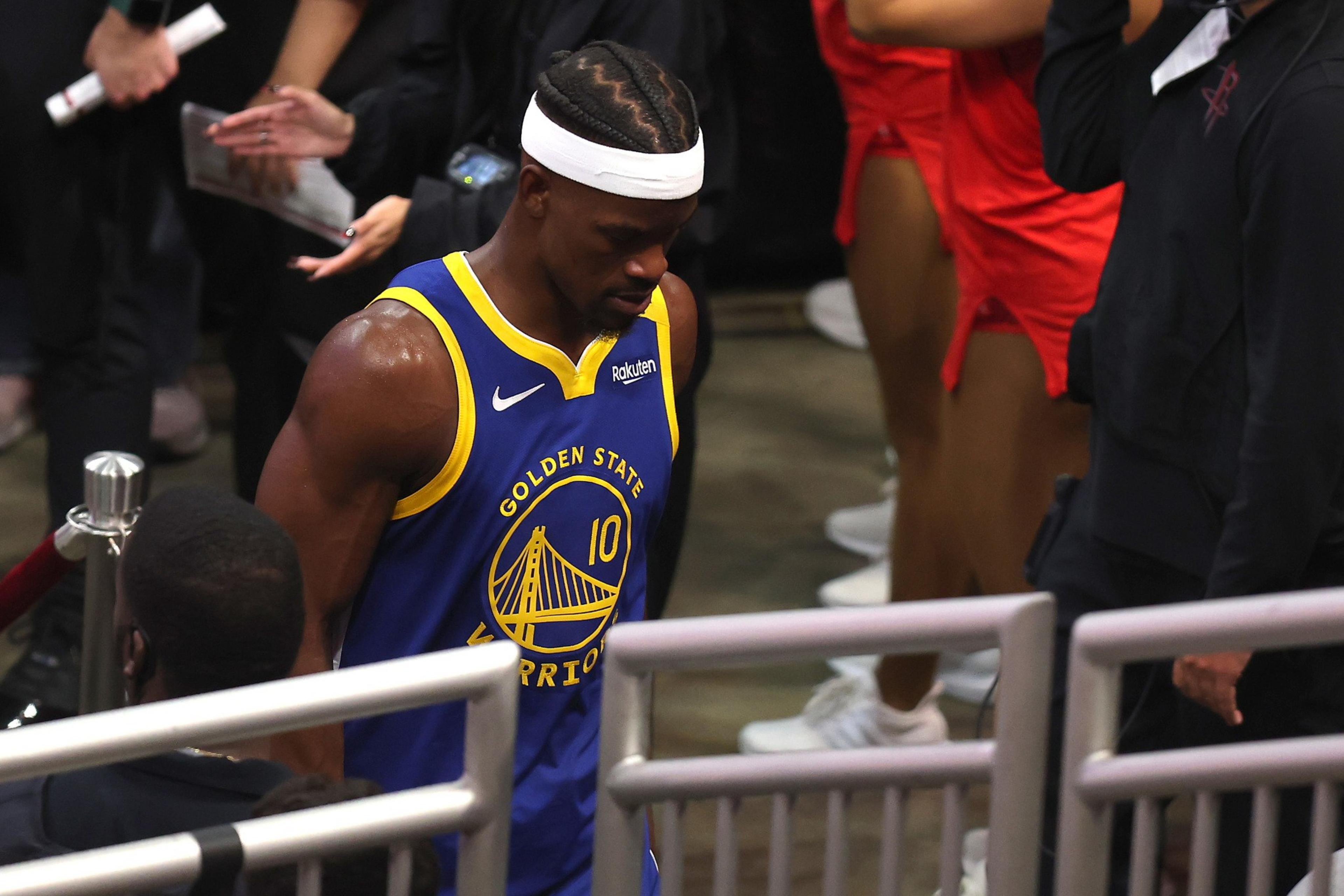 A basketball player in a blue &quot;Golden State Warriors&quot; jersey with the number 10 walks through a crowd. He wears a white headband and has braided hair.