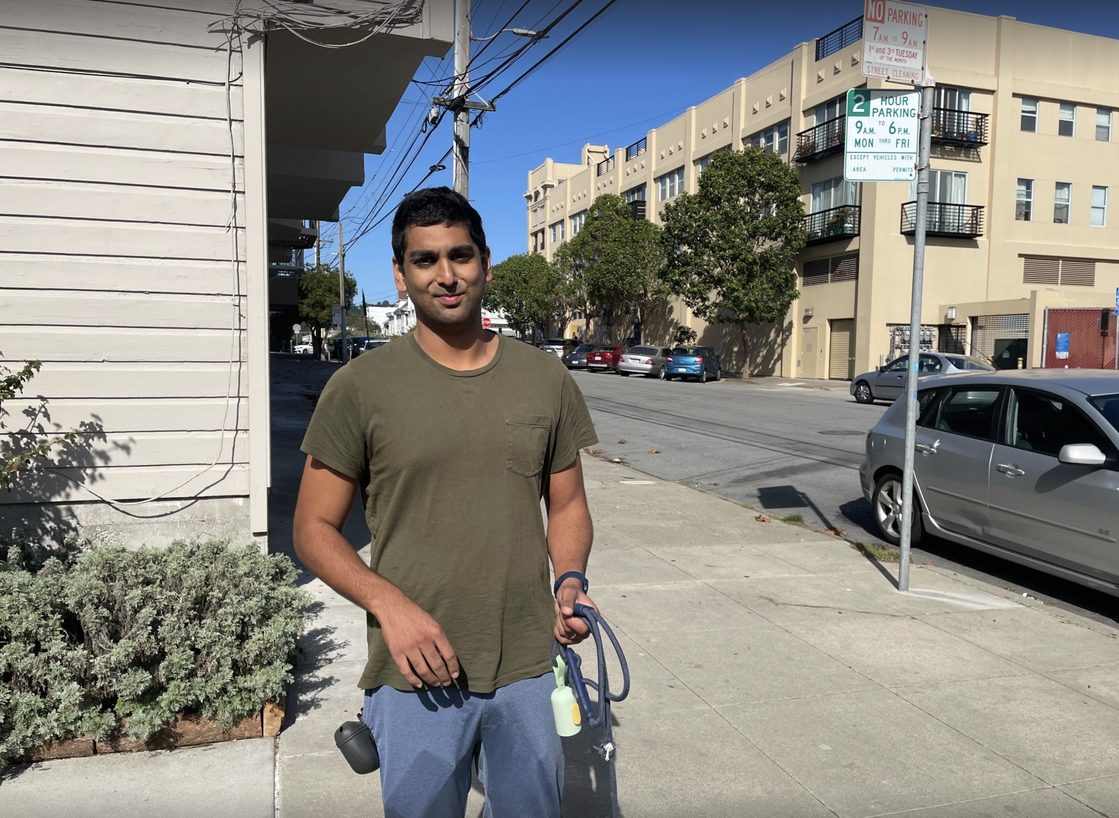 A man in a green t-shirt and blue pants stands on a sidewalk holding a leash and a small bottle, with buildings and parked cars behind him.
