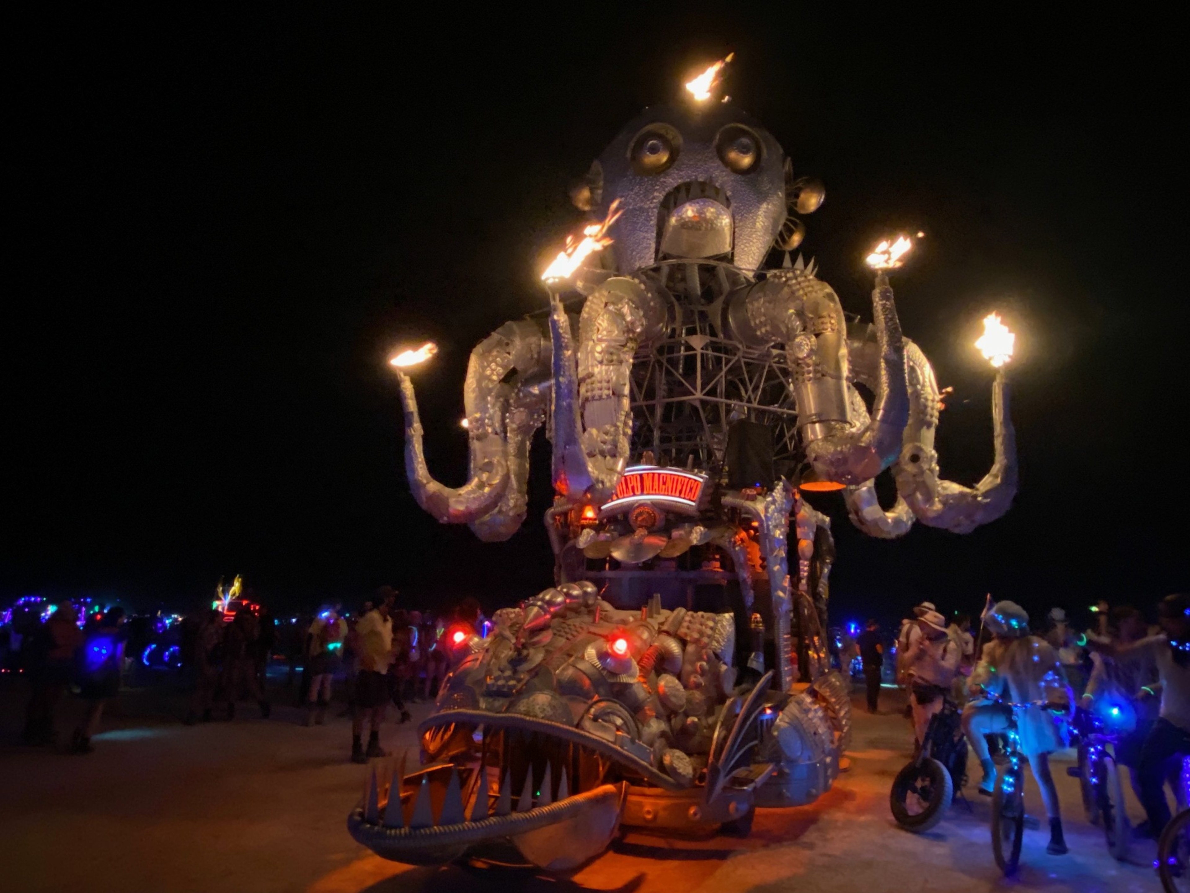 An art car shaped like a mechanical octopus with flaming tentacles illuminates a nighttime desert event, surrounded by colorful lights and people biking.