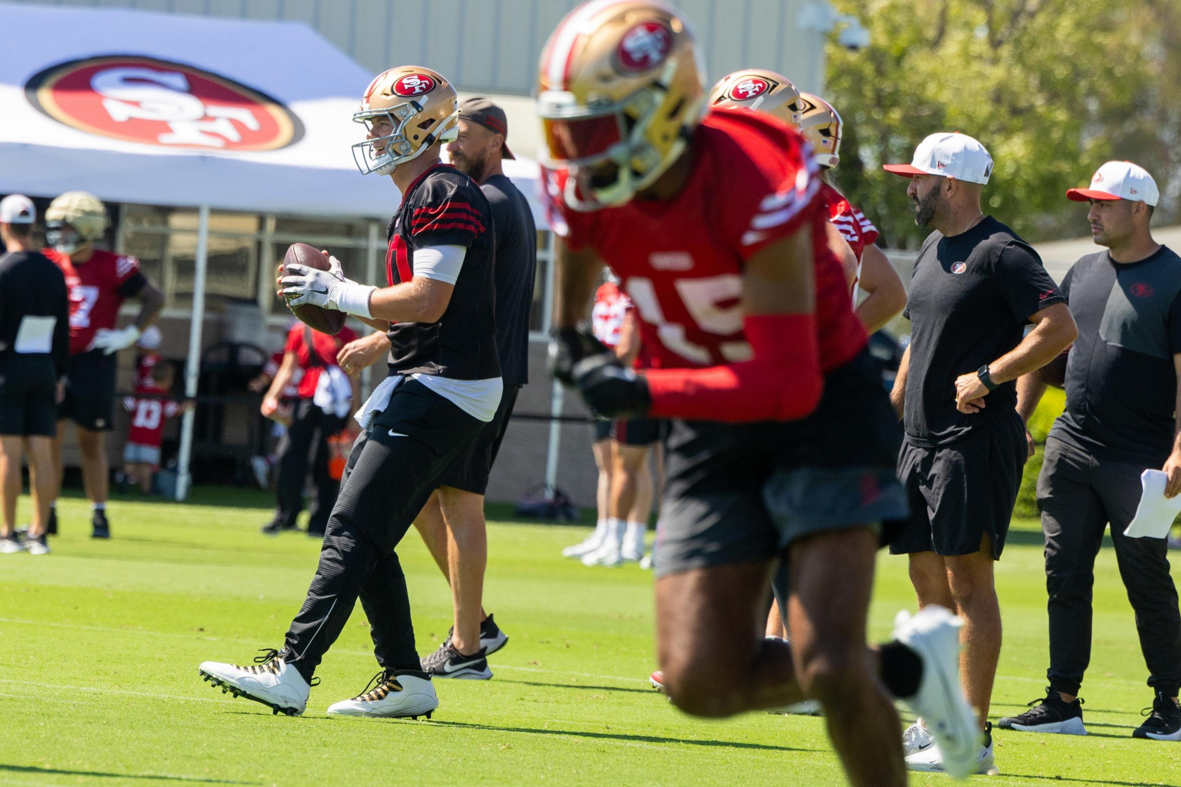 Football players in red and black gear practice on a field, with coaches observing. One player holds a ball, others run, all wearing helmets with a logo.