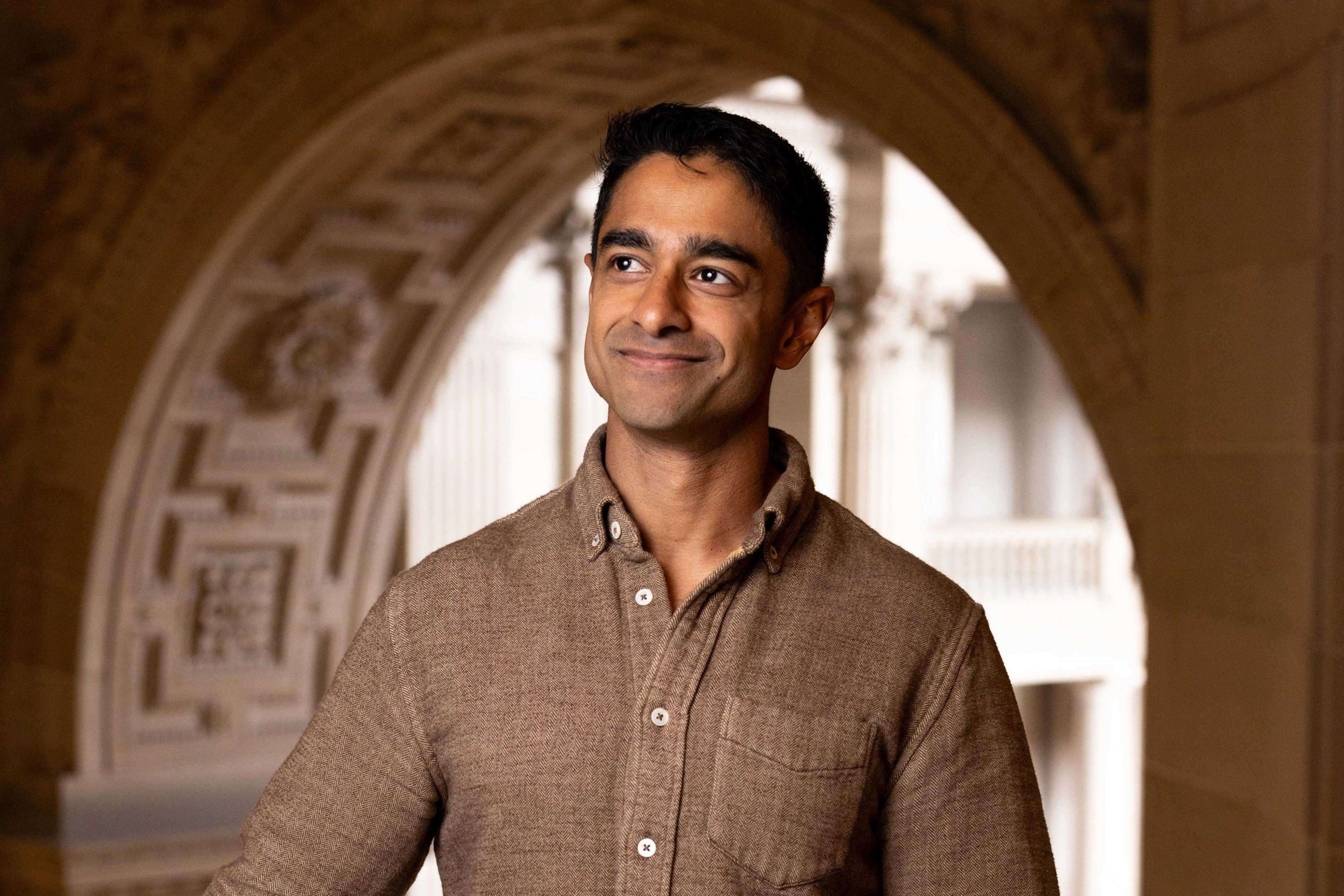 A person stands indoors, wearing a brown collared shirt and smiling slightly. The background features ornate architectural details and soft lighting.
