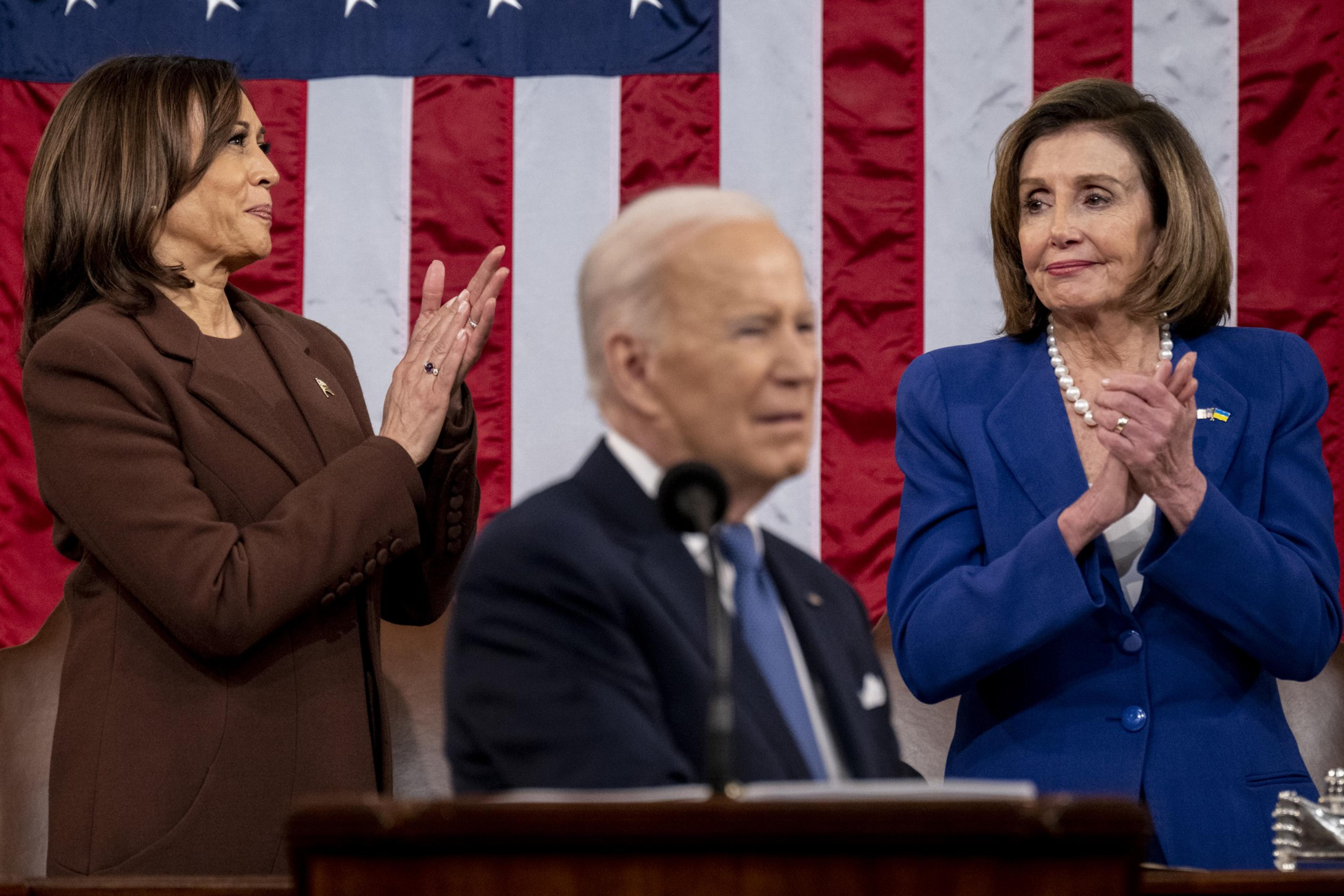 A blurred man at a podium is flanked by two women in brown and blue suits, clapping, with an American flag as the backdrop.