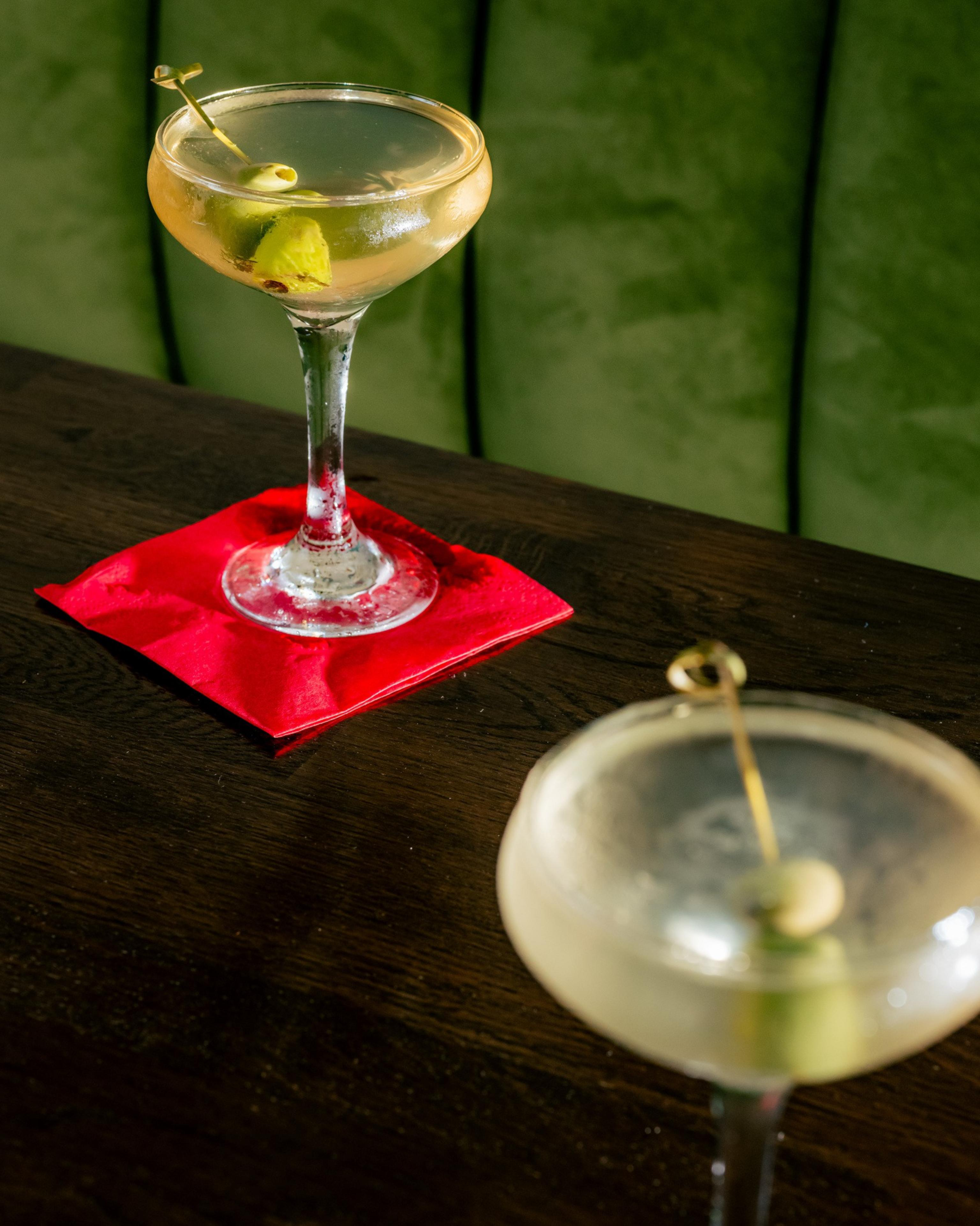 Two martini glasses sit on a dark wooden table. One is on a red napkin, both garnished with olives. The background is a green upholstered surface.