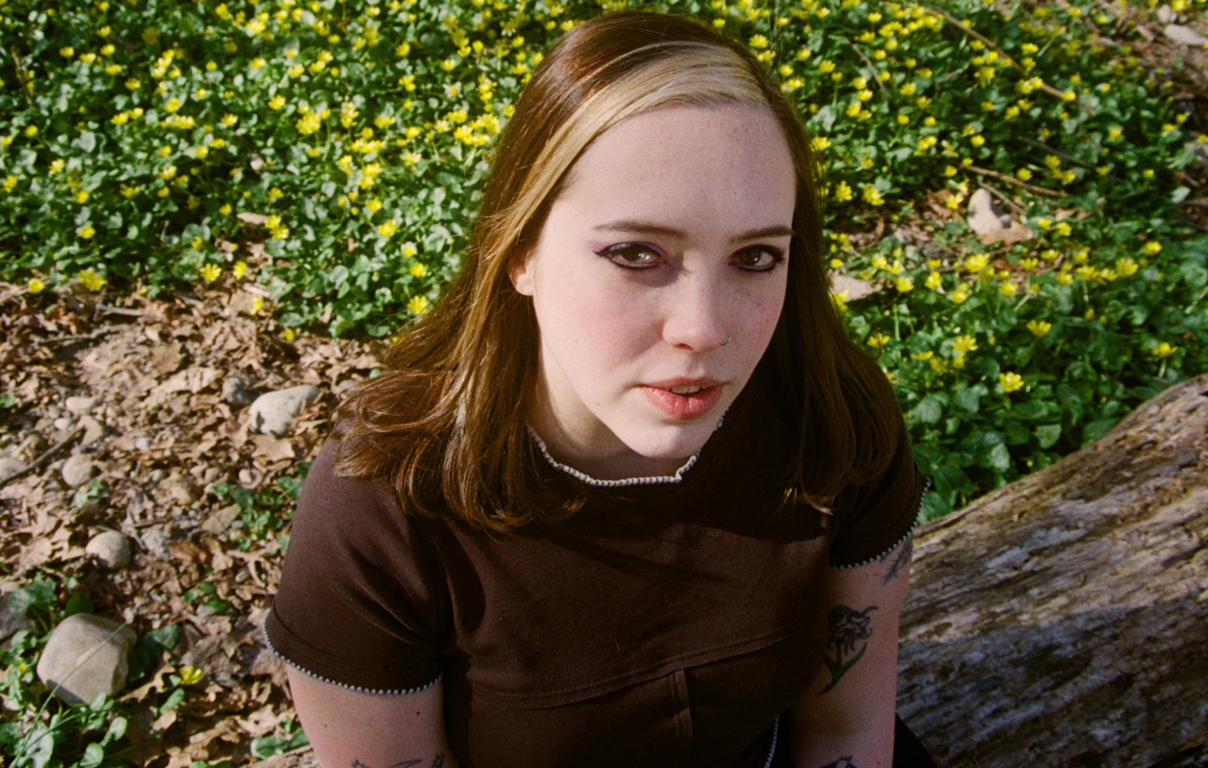 A person with long hair, partially dyed blonde, is sitting outdoors on a log. They are wearing a dark shirt with tattoos visible on their arms. Yellow flowers bloom in the background.