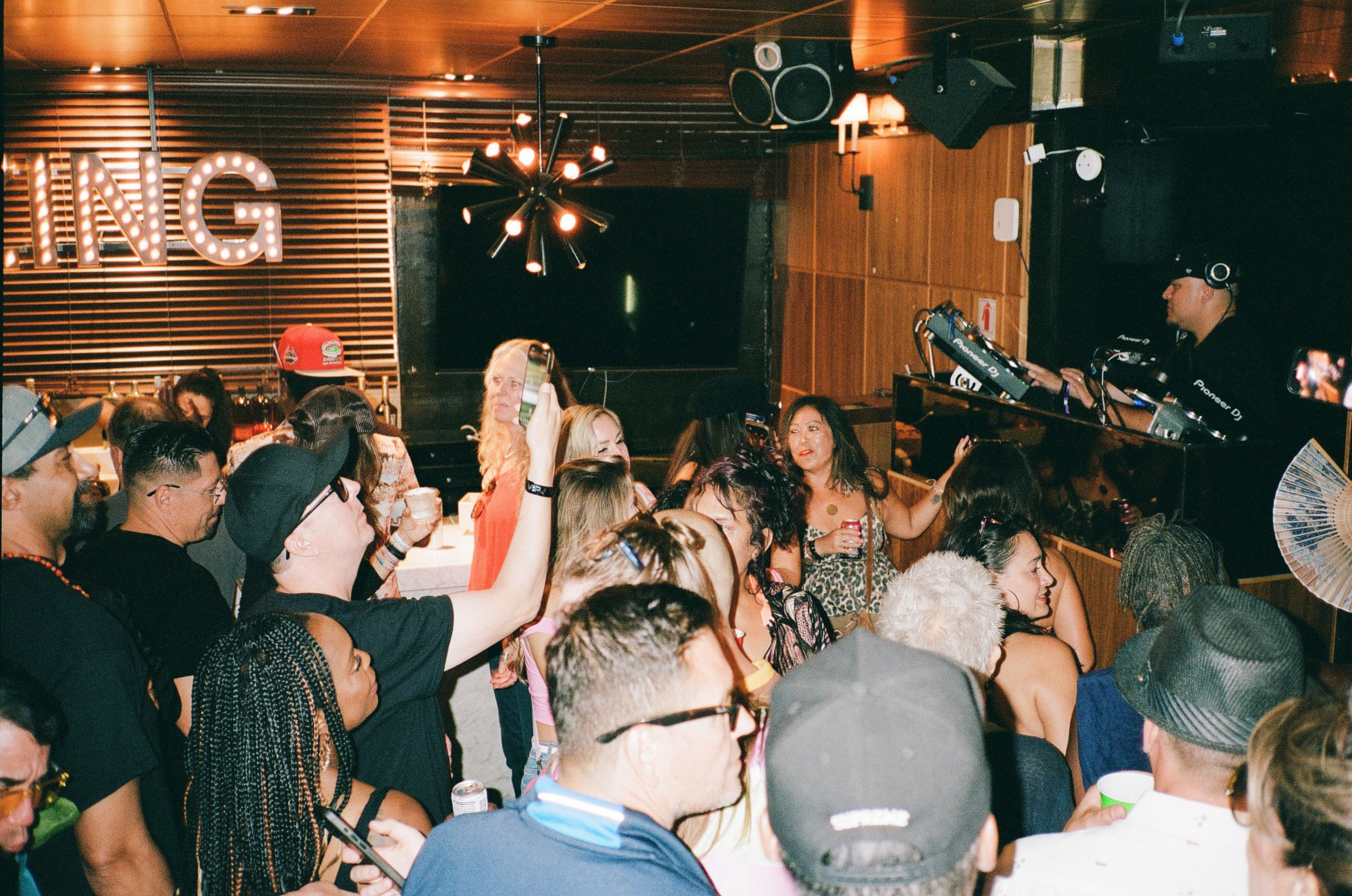 A crowded indoor party with people drinking, dancing, and a DJ playing music in a warmly lit room with a sign partially reading "ING."