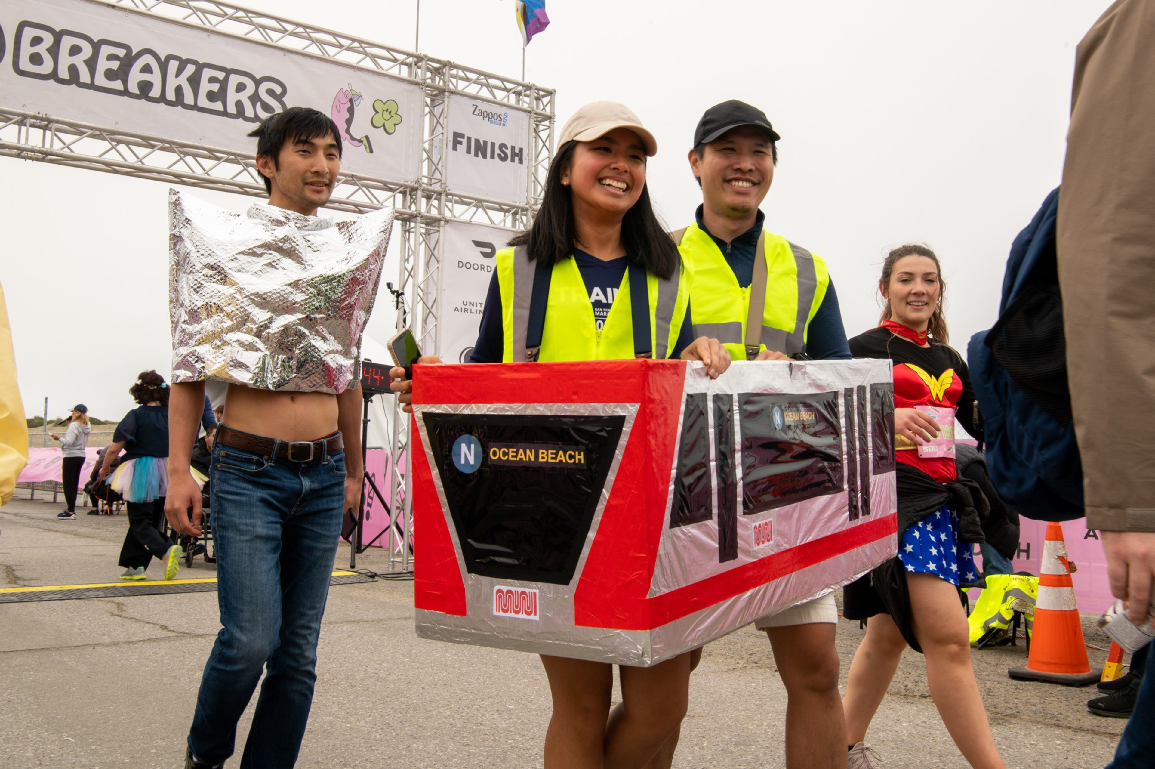 Three people walk near a finish line; two wear a subway train costume labeled "Ocean Beach," one wears a shiny silver foil outfit, and another dresses as Wonder Woman.