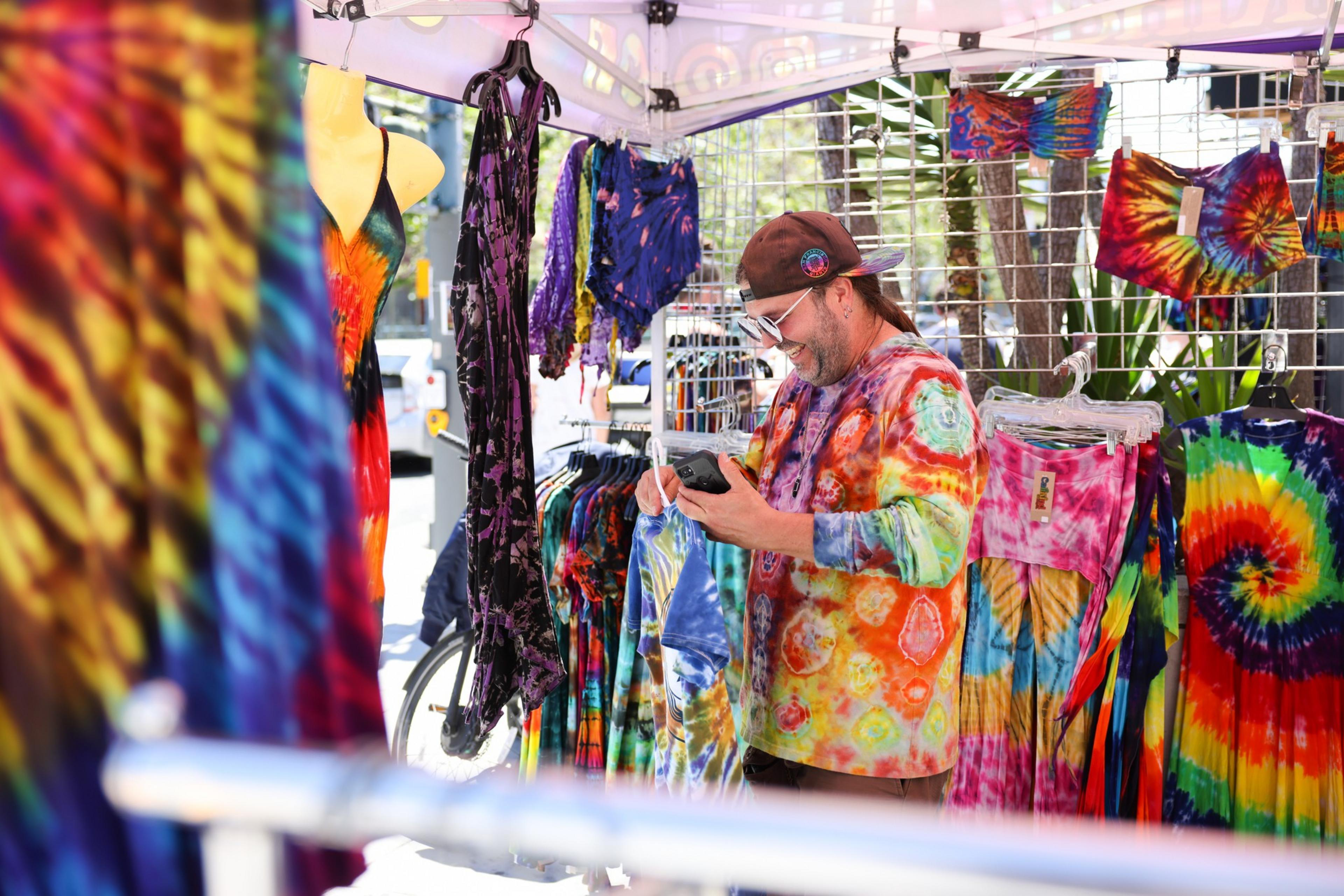 A man, smiling and wearing a tie-dye shirt, stands under a tent filled with colorful tie-dye clothing. He looks at a phone, surrounded by vibrant garments.