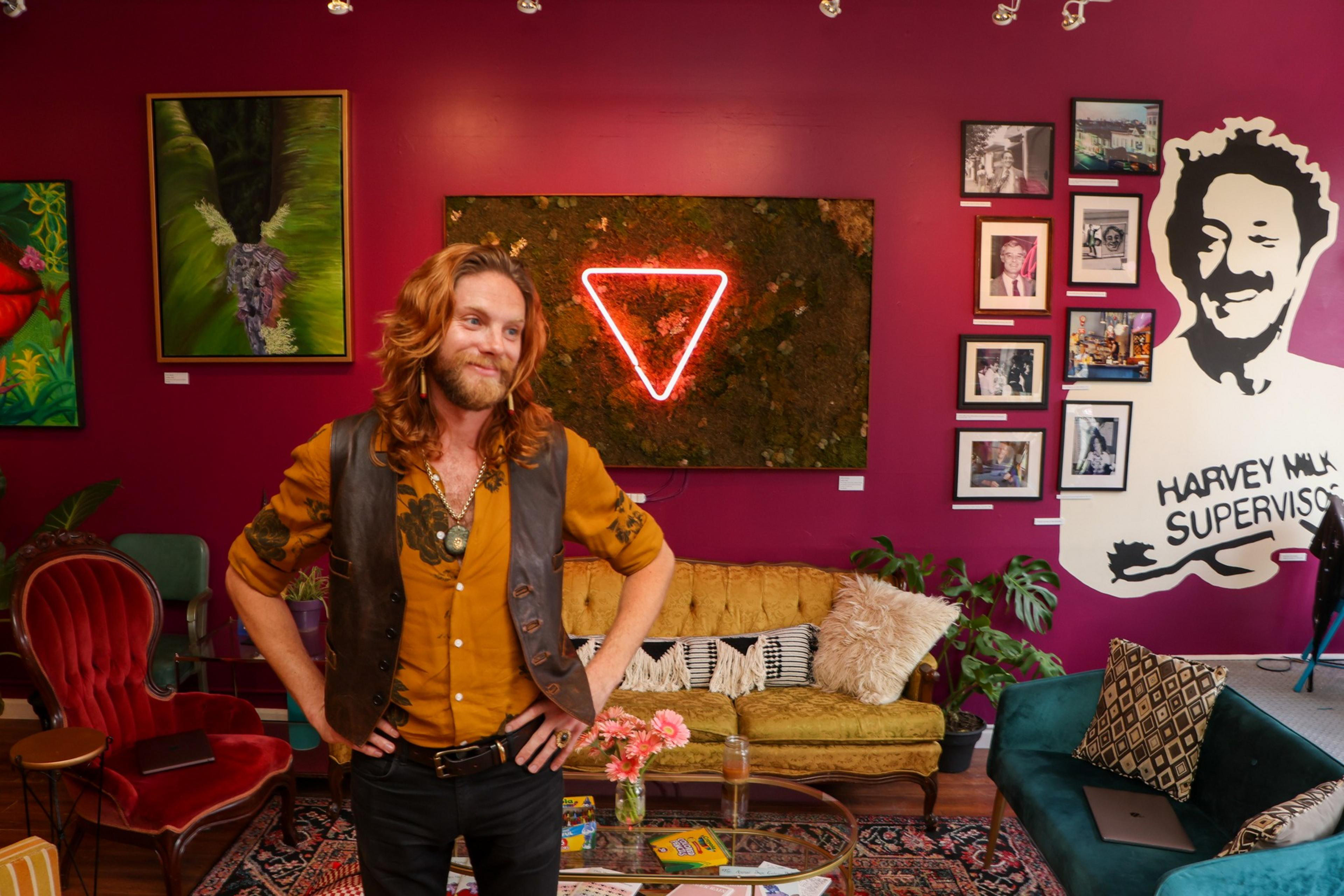 A man with long hair and a beard stands smiling in a colorful room with vintage furniture and wall art, including a neon triangle and a Harvey Milk mural.