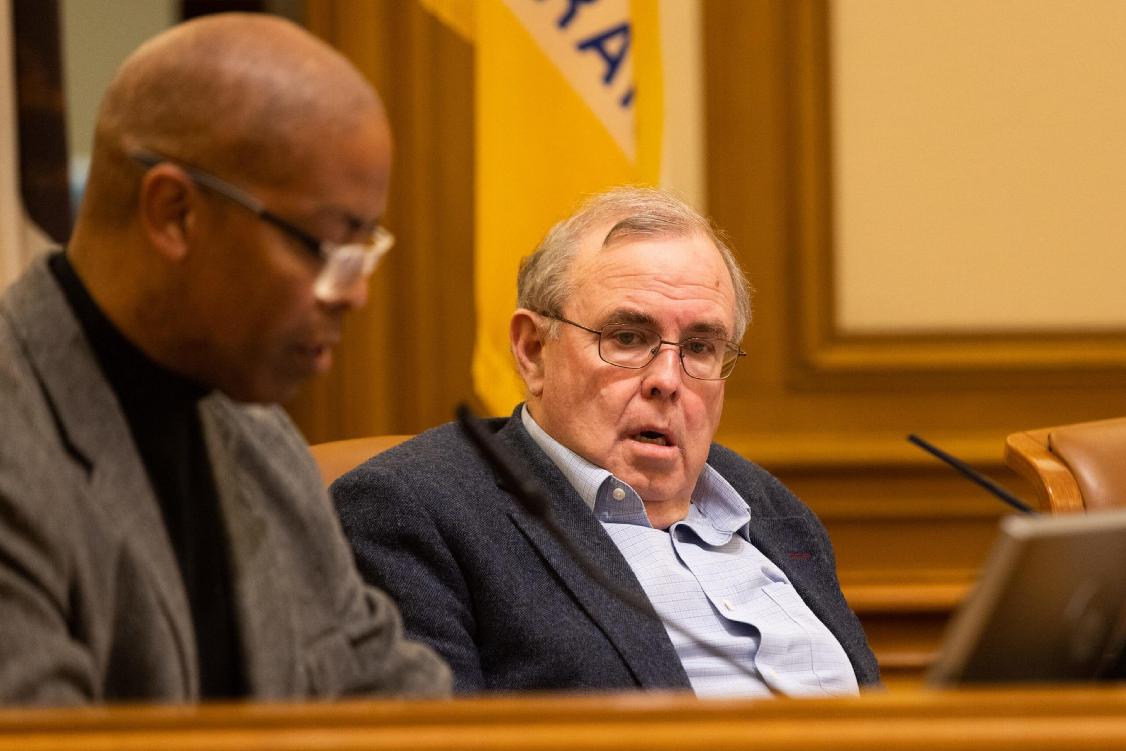 An elderly man with glasses and a blue blazer looks surprised while sitting next to a bald man in glasses and a gray jacket, both at a wooden desk.