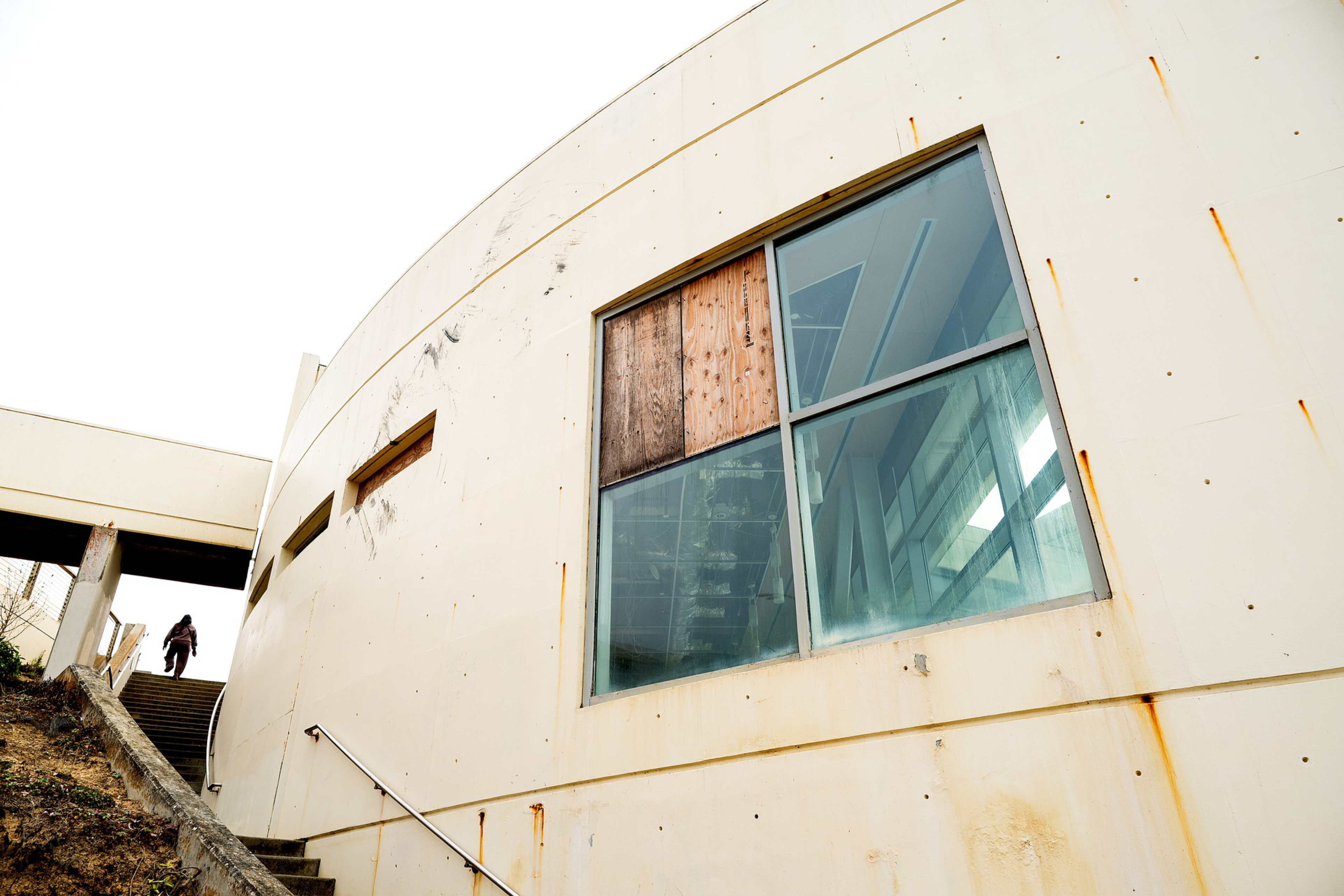 A large, curved building wall with rust stains and a window partially boarded up with wood, stairs leading up to an entrance where a person is walking.