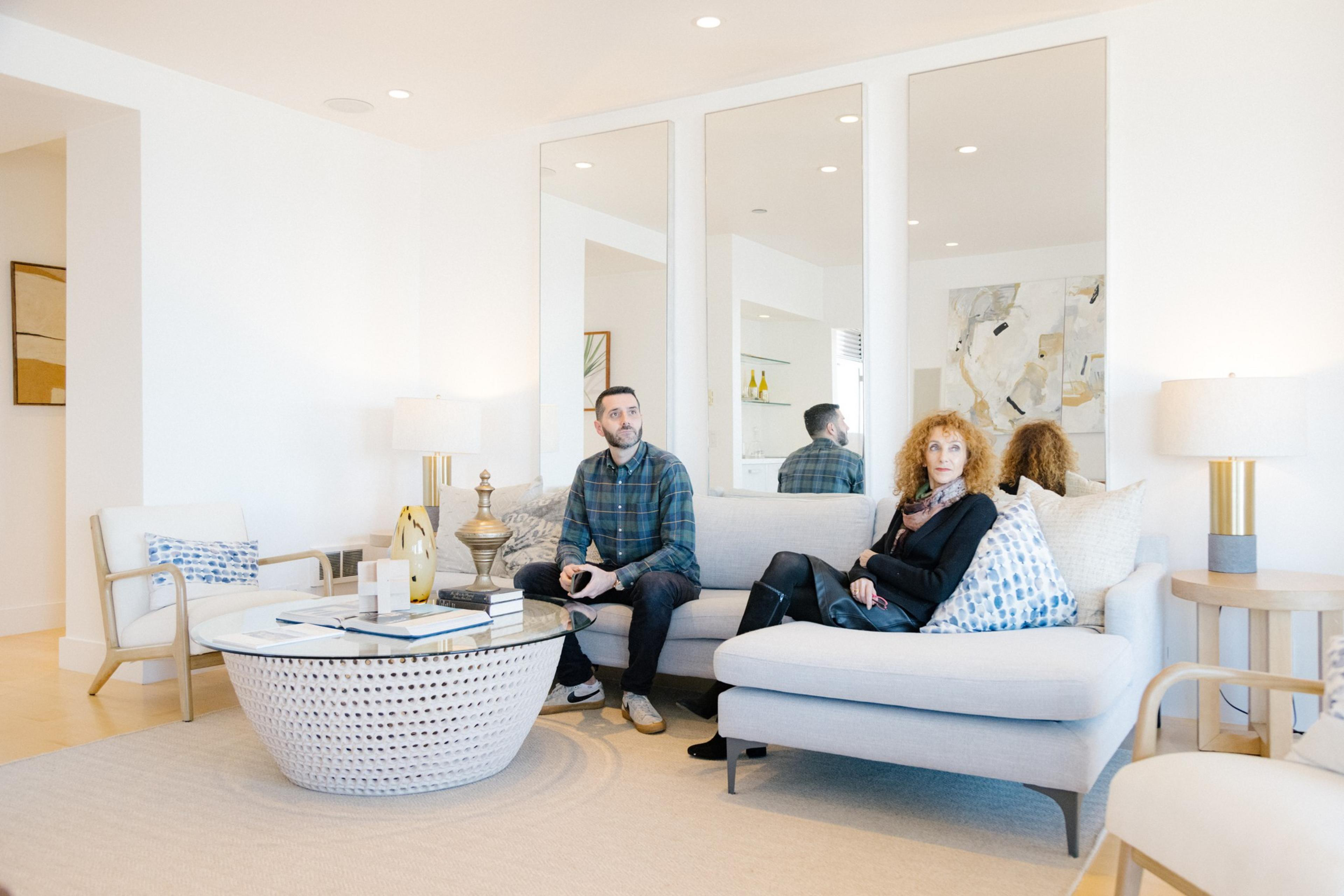 A bright living room with two people sitting on a couch, modern decor, ample lighting, and large mirrors on the wall.