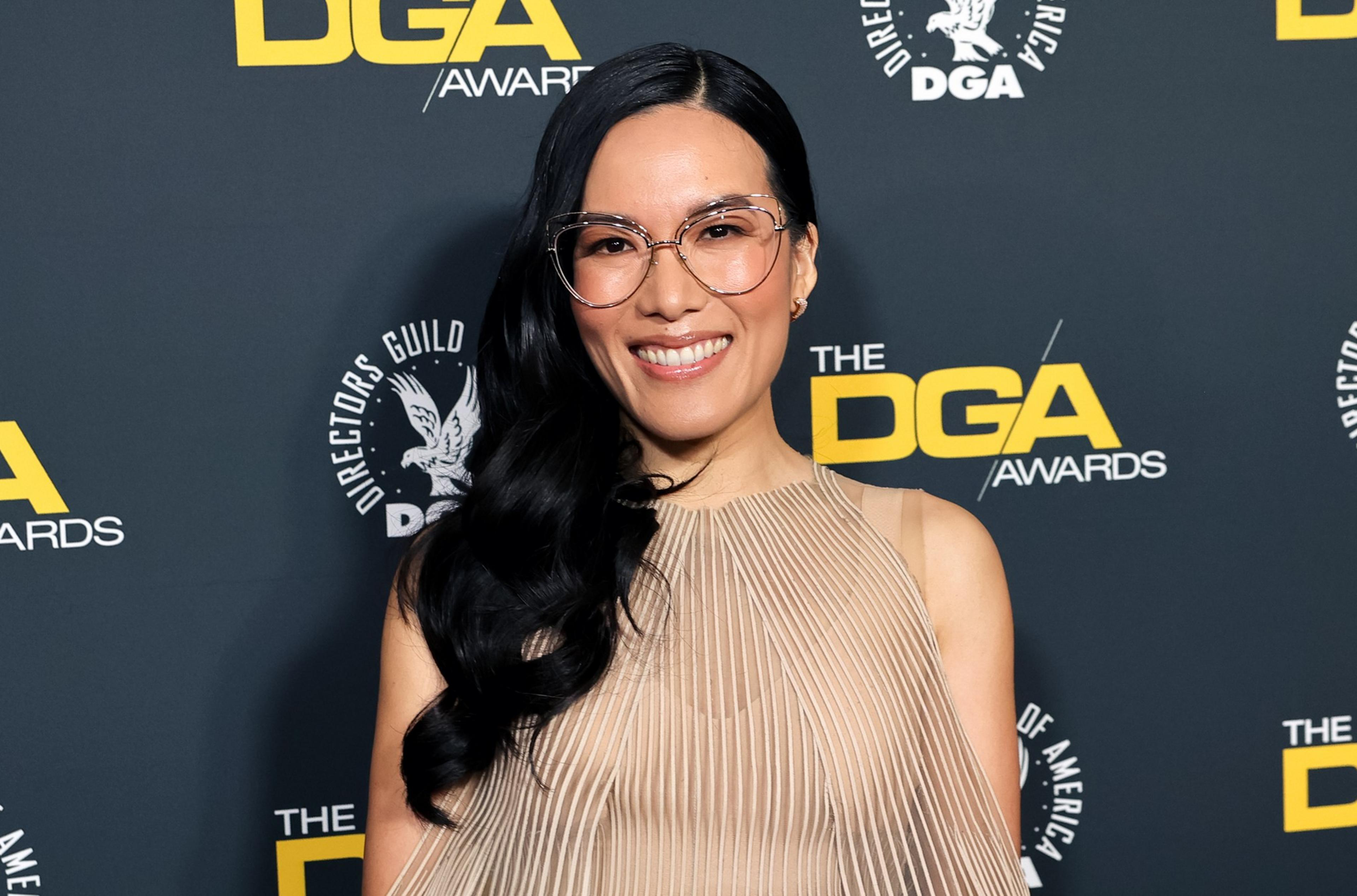 A person with long dark hair, wearing glasses and a beige dress, smiles against a backdrop featuring yellow "DGA Awards" text and logos.