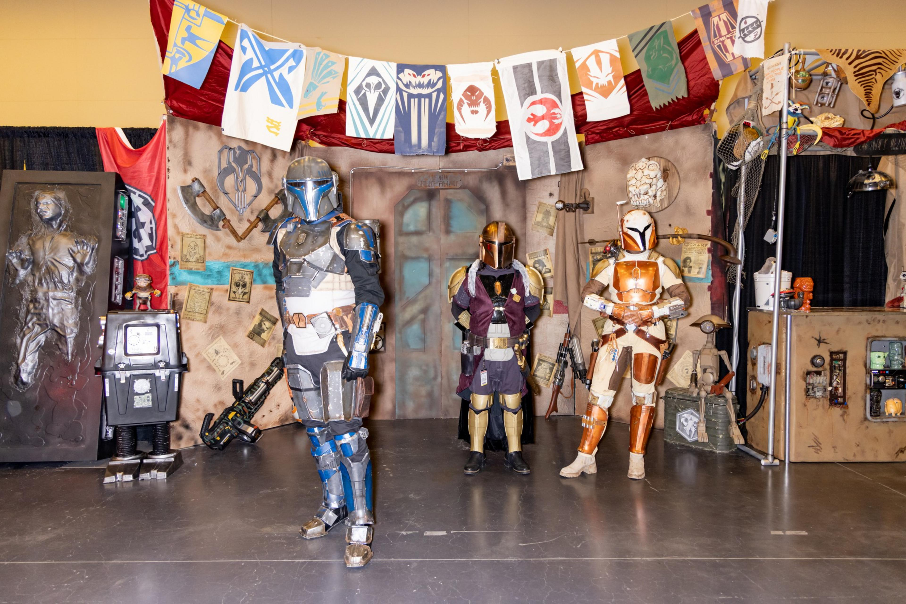 Three people in Mandalorian armor pose in a sci-fi-themed room with a decorated backdrop and banners. Props and a figure encased in carbonite are also visible.