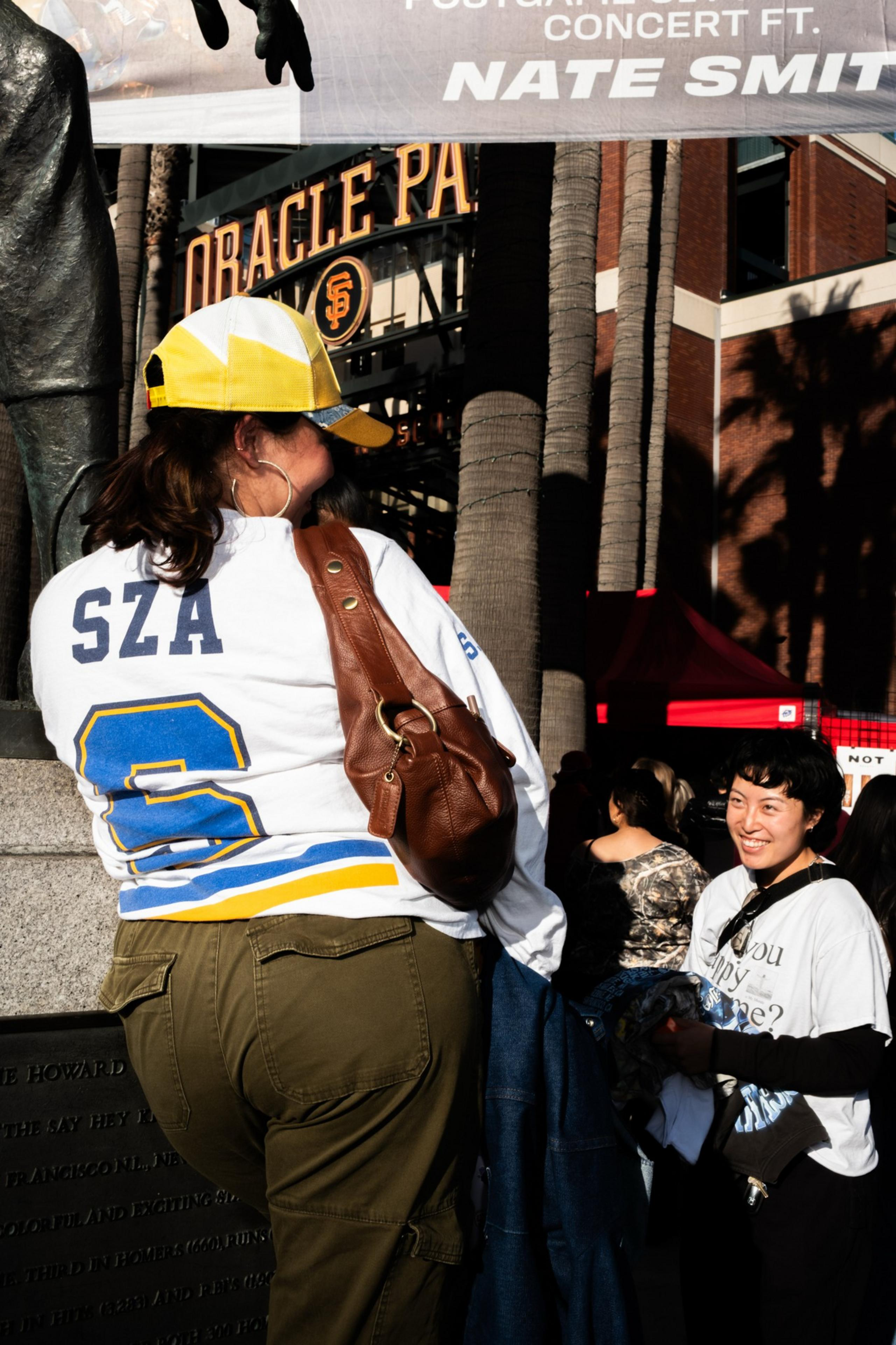 A person wearing a yellow cap and a white shirt with &quot;SZA&quot; and a number stands near a statue at Oracle Park, chatting with another smiling person.