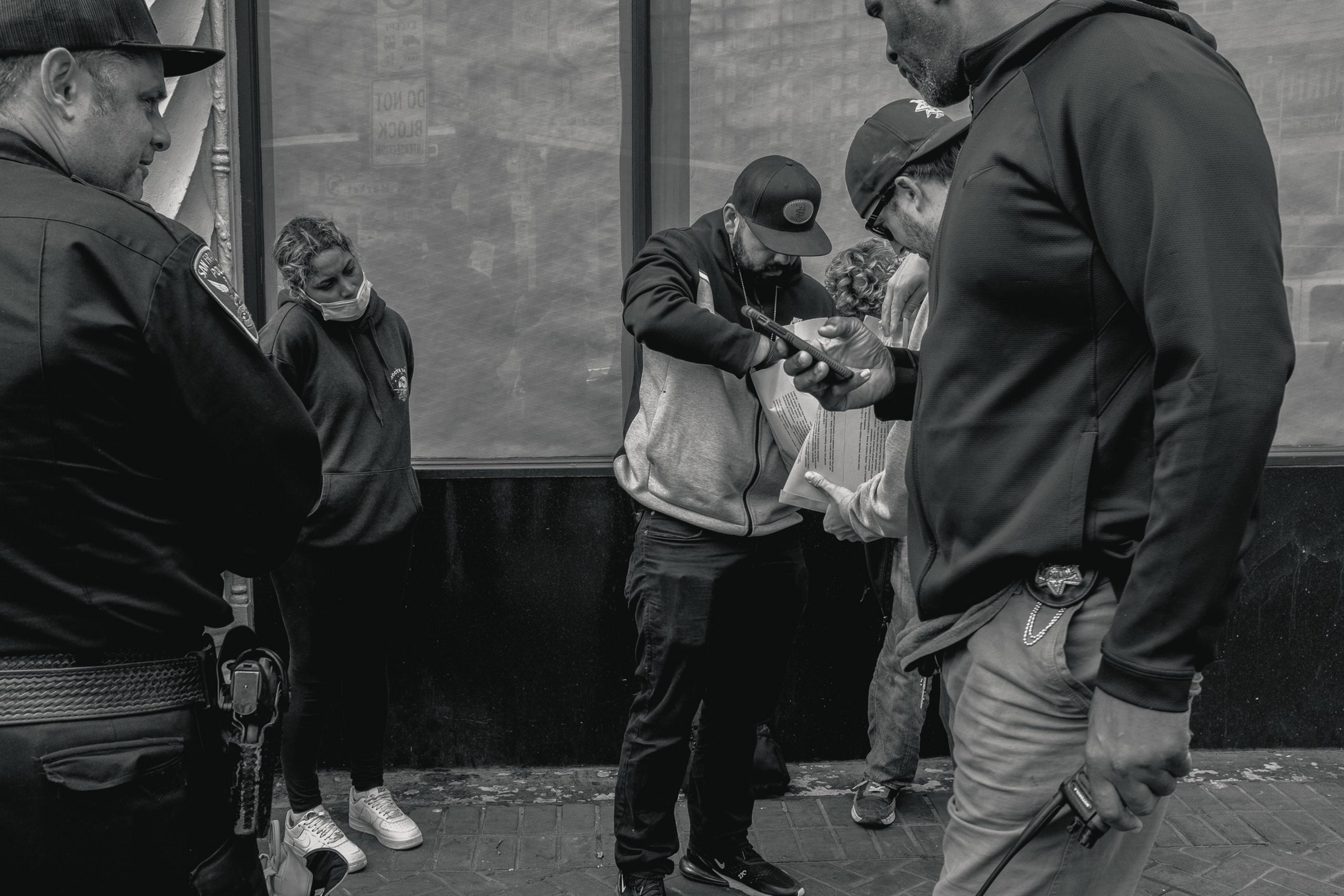 A group of people stand on a sidewalk, with one person appearing to show something on a phone to another. A masked woman leans against the wall nearby.