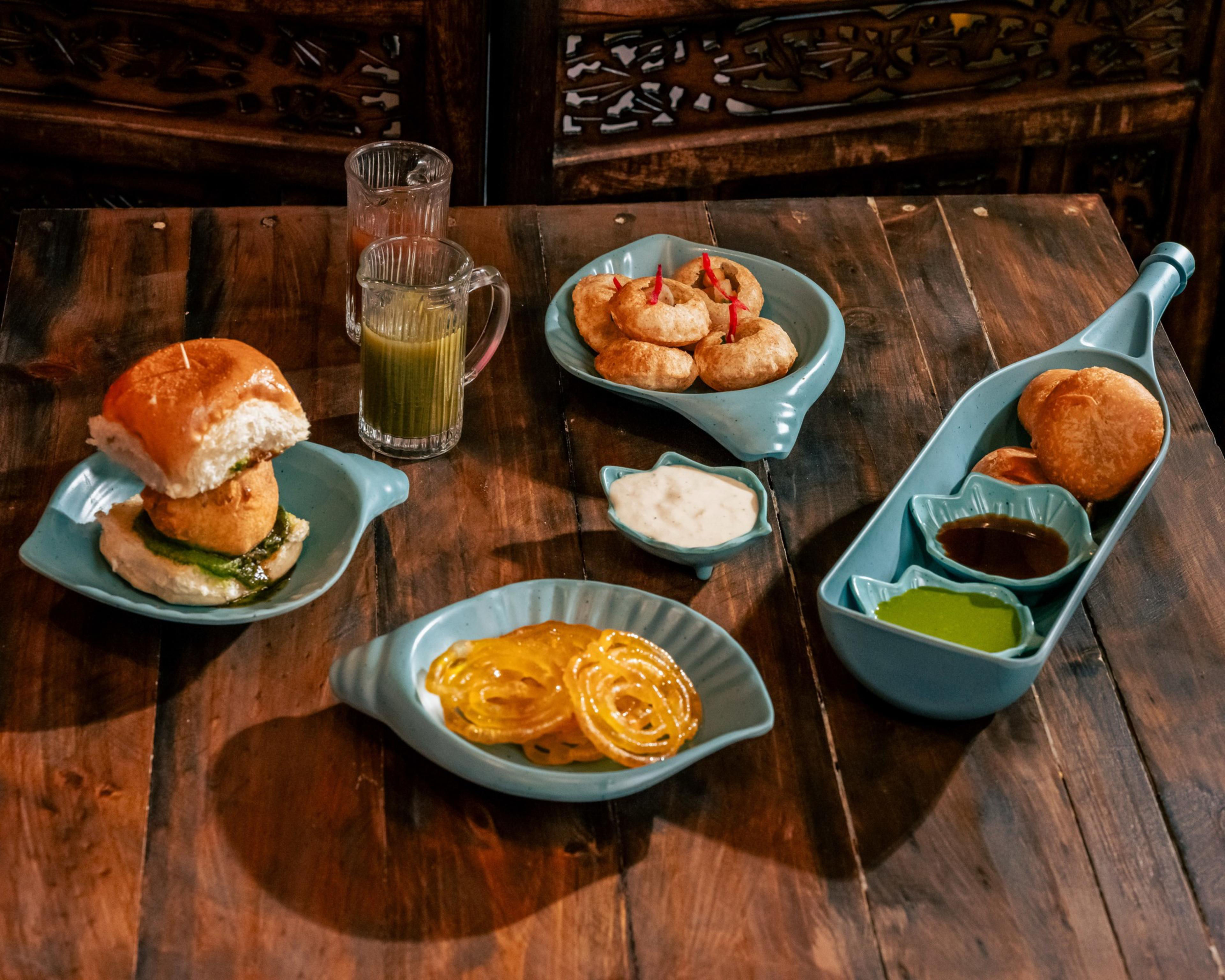 A wooden table holds plates of Indian snacks including vada pav, pani puri, jalebi, and chutneys, with two glasses of green and pink drinks.