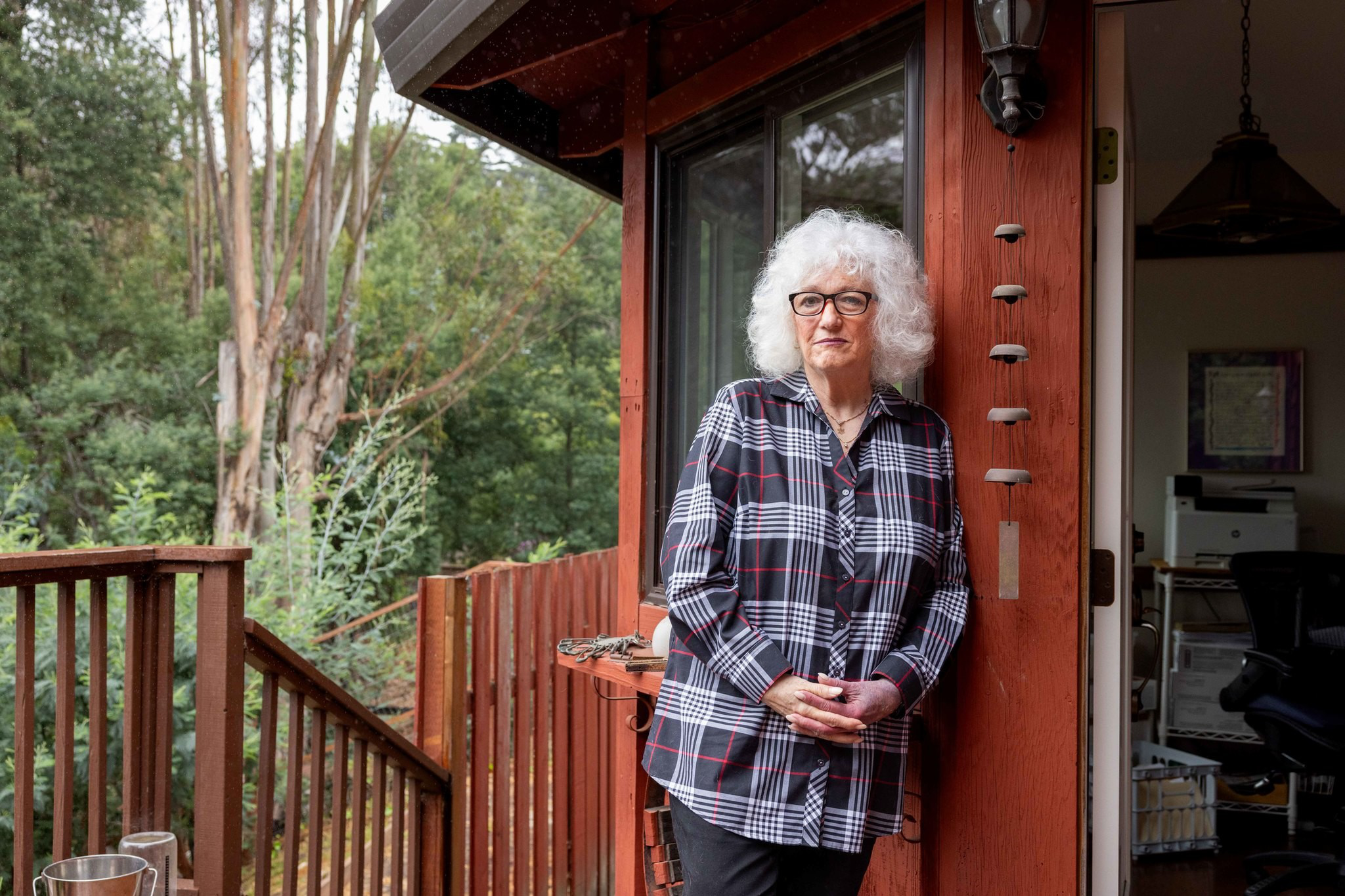 An elderly woman with curly white hair and glasses stands on a wooden porch, leaning against a red wall outside a house surrounded by trees.