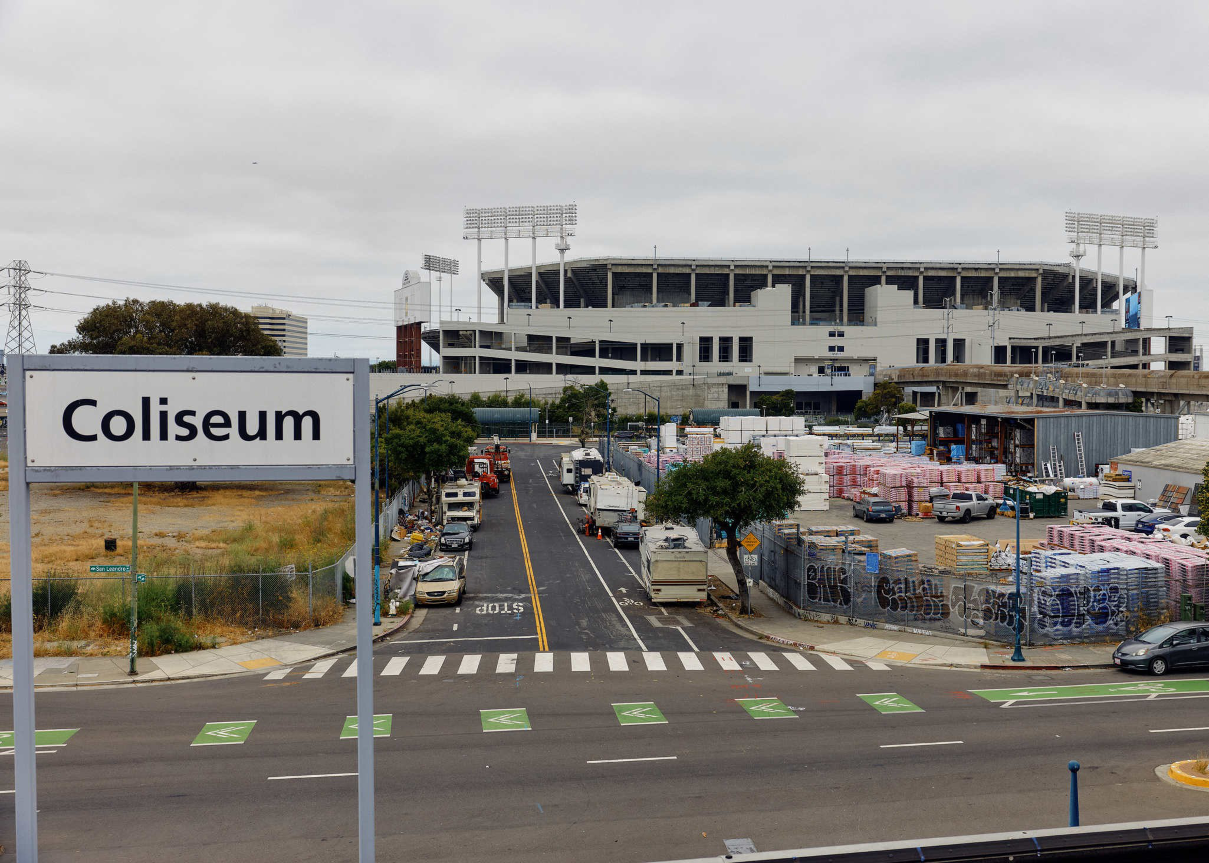A large stadium labeled “Coliseum” stands behind a street lined with parked vehicles and industrial materials under an overcast sky.