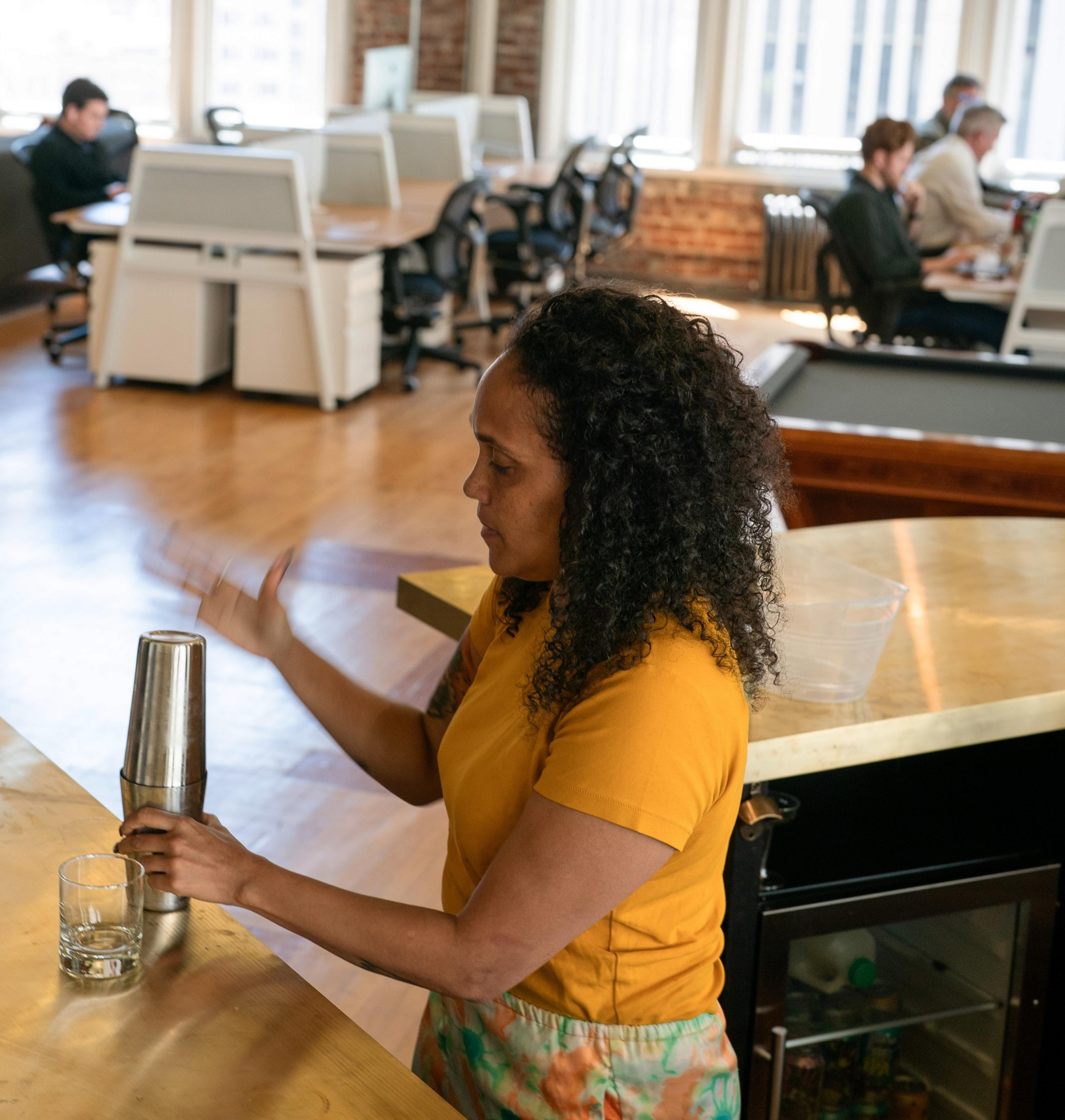 A woman in a yellow shirt makes a drink with a shaker in an office kitchen, while several people work at desks in the background.