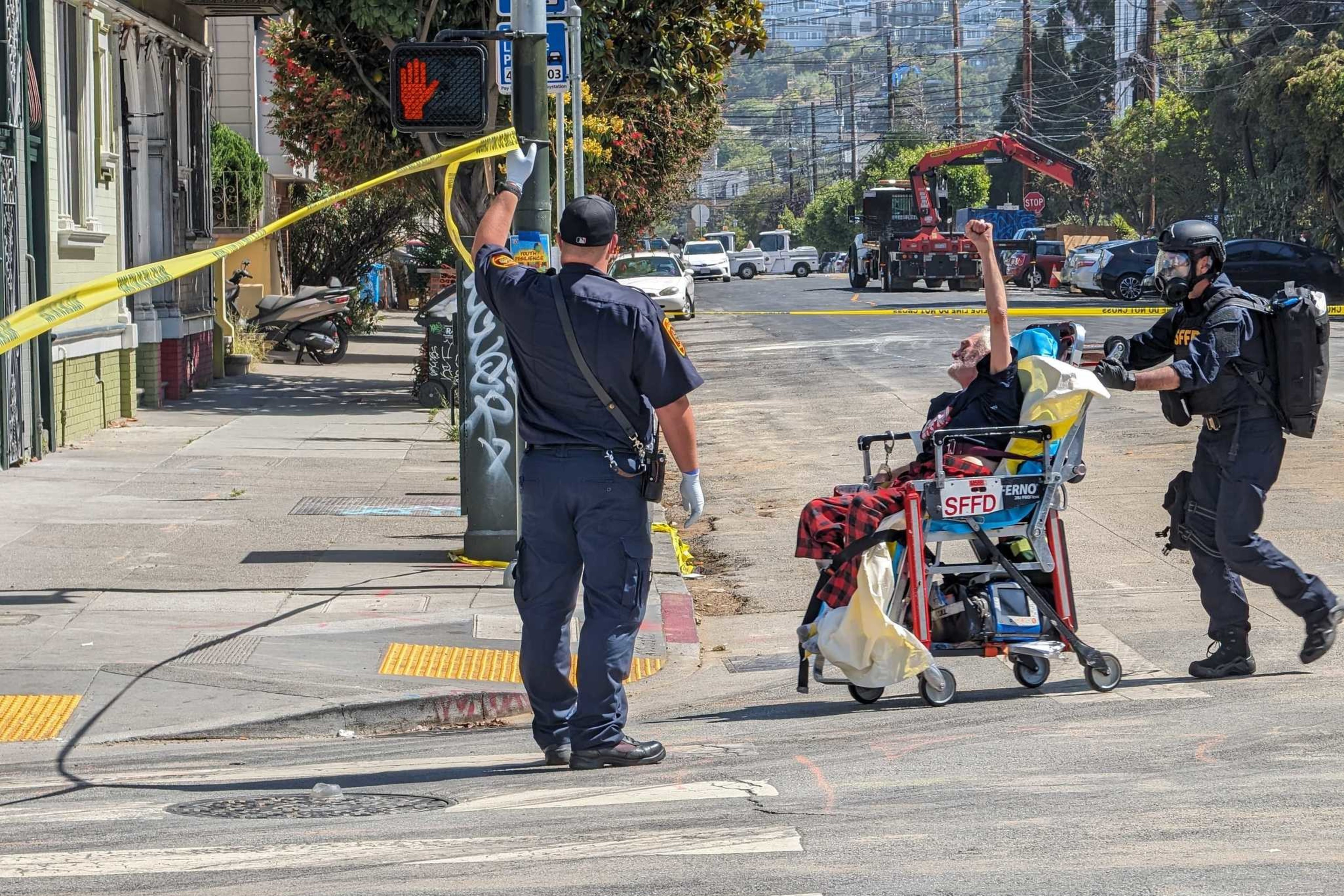 A person in a wheelchair is escorted by two officers, one holding a roll of caution tape. The street is cordoned off, with a "Do Not Walk" signal visible.