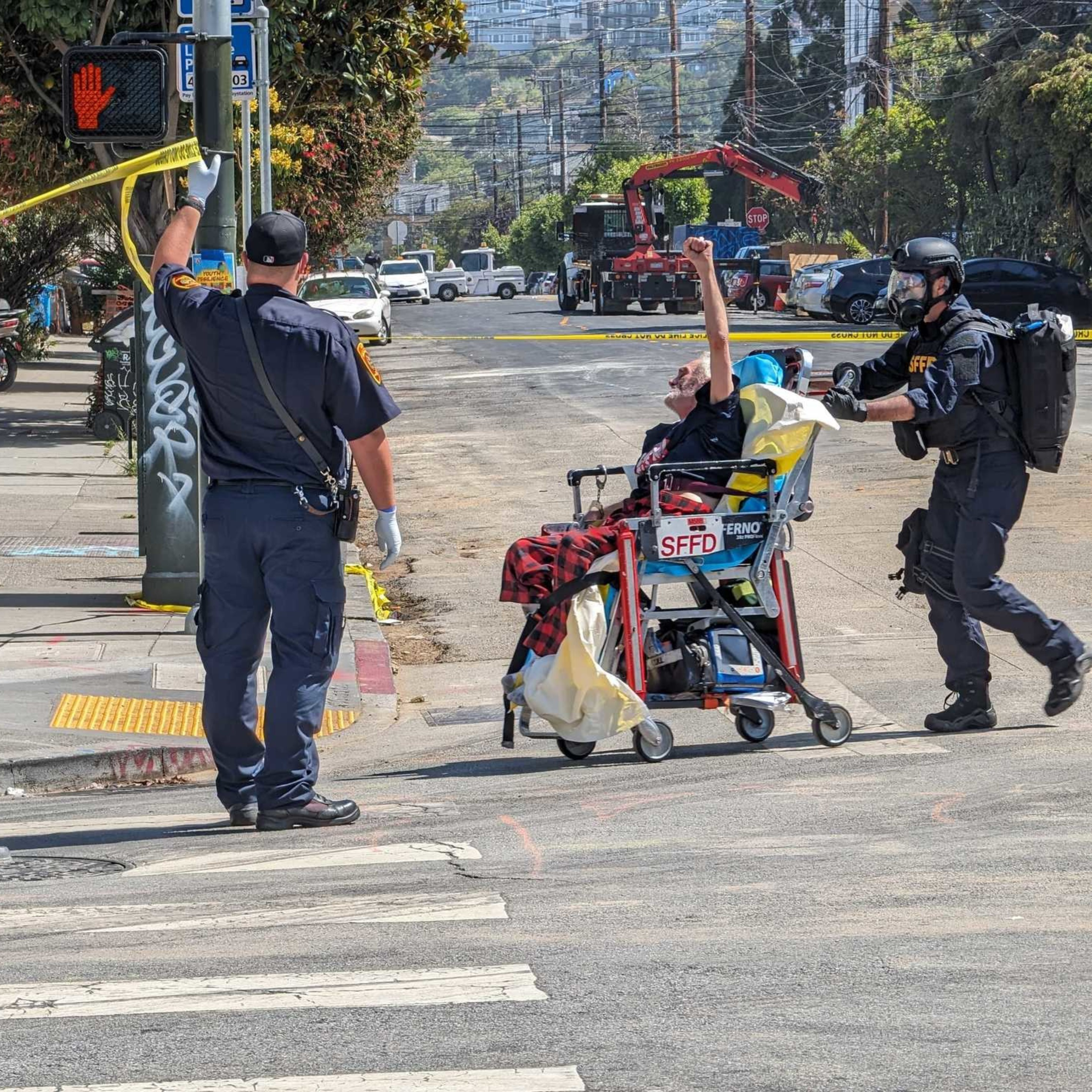 A person in a wheelchair is escorted by two officers, one holding a roll of caution tape. The street is cordoned off, with a "Do Not Walk" signal visible.