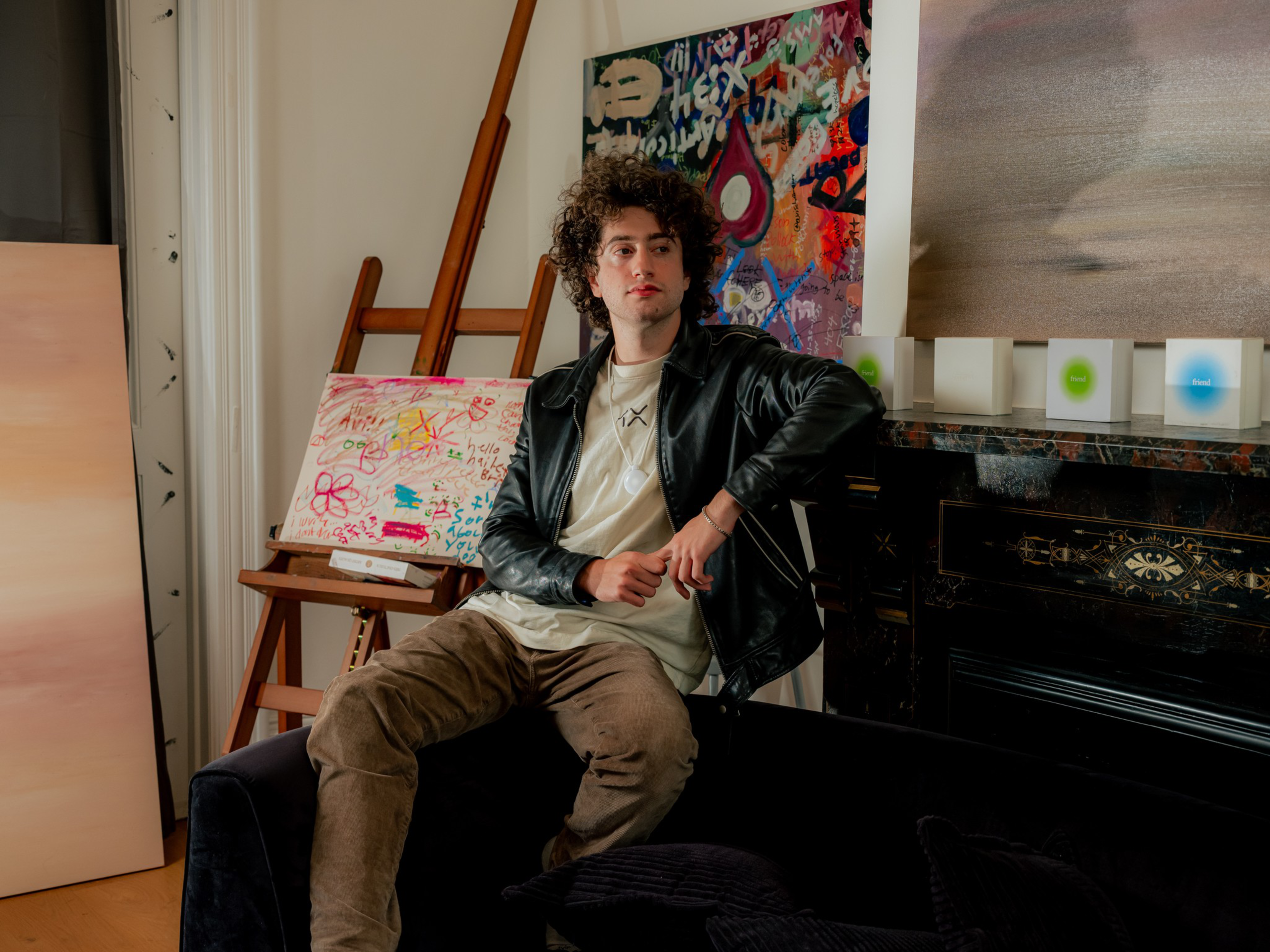 A young person with curly hair sits on a dark couch, wearing a black leather jacket and beige pants, with colorful abstract paintings and an easel in the background.