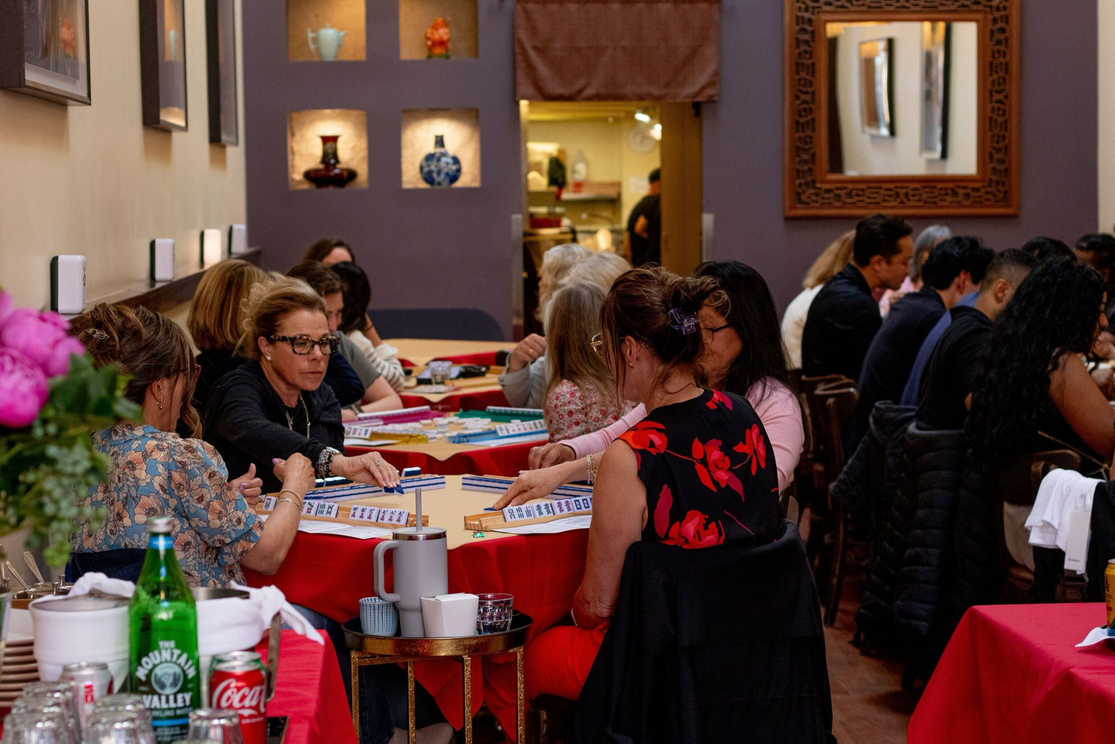Several groups of people sit at red-covered tables inside a warmly lit room, engaged in playing Mahjong, with decorative vases on shelves in the background.