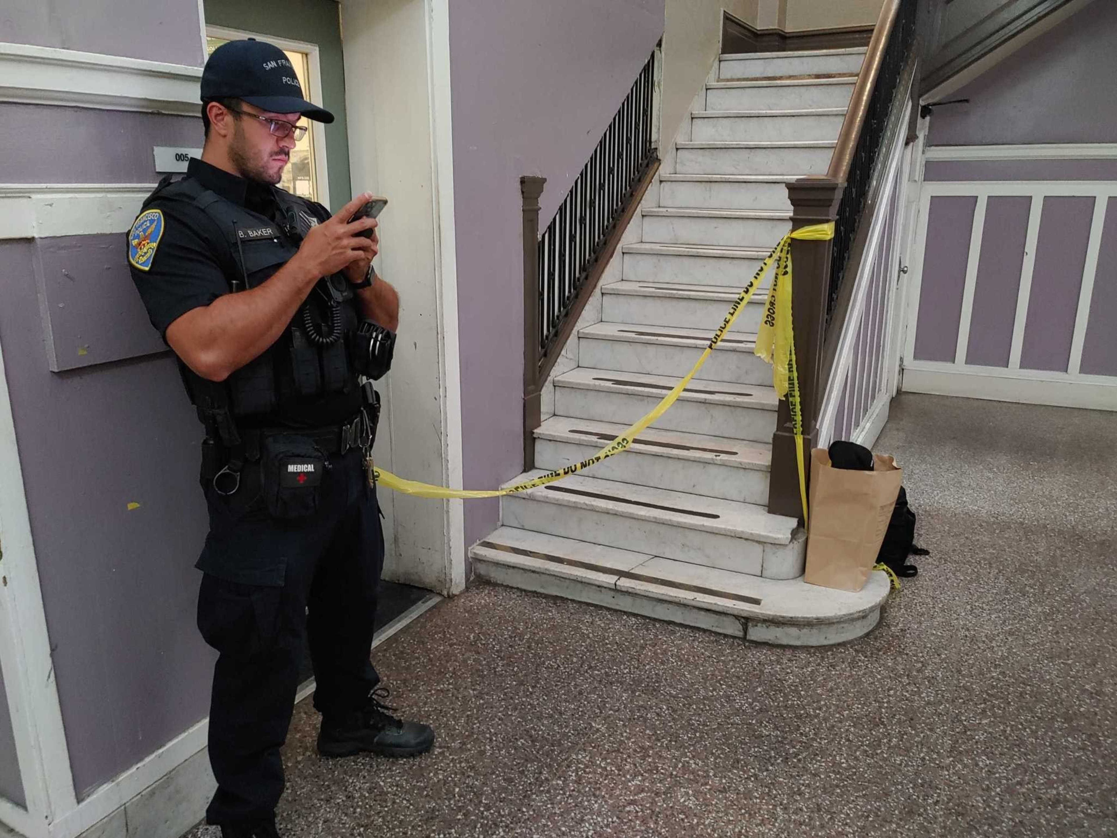 A police officer stands by yellow caution tape blocking off a stairwell, while looking at a phone, with a paper bag and a black backpack at the stairs.