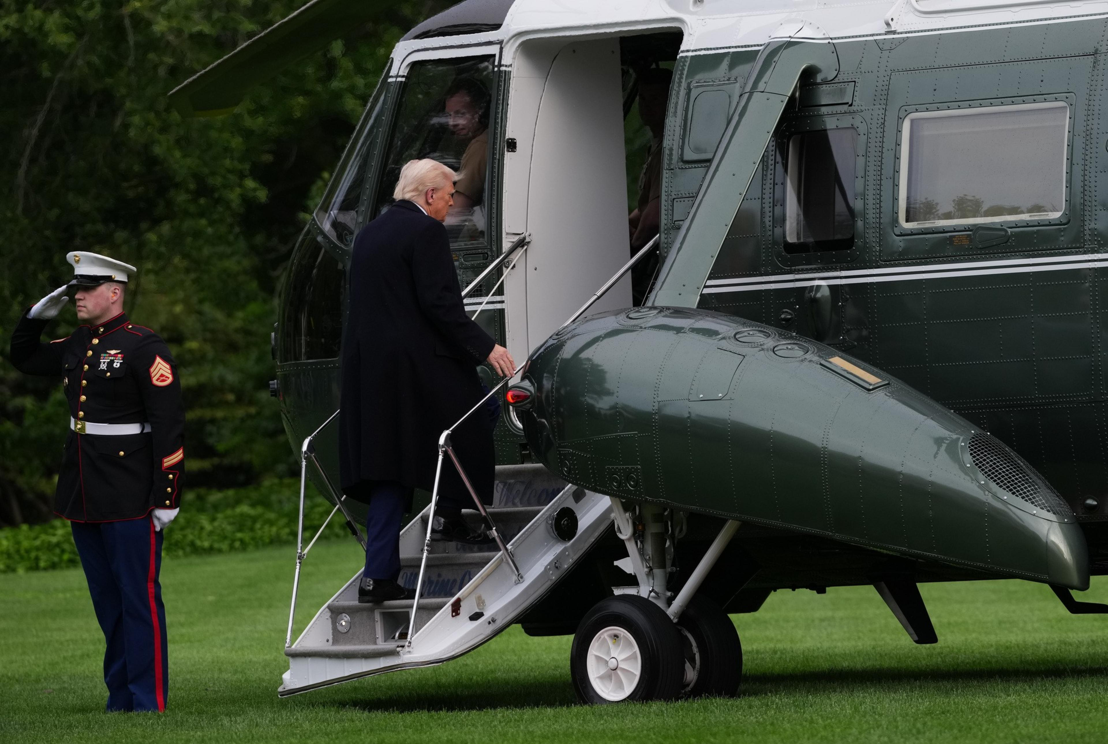 A man in a dark coat is boarding a green and white helicopter. A uniformed Marine stands saluting nearby. The scene is set on a grassy area.