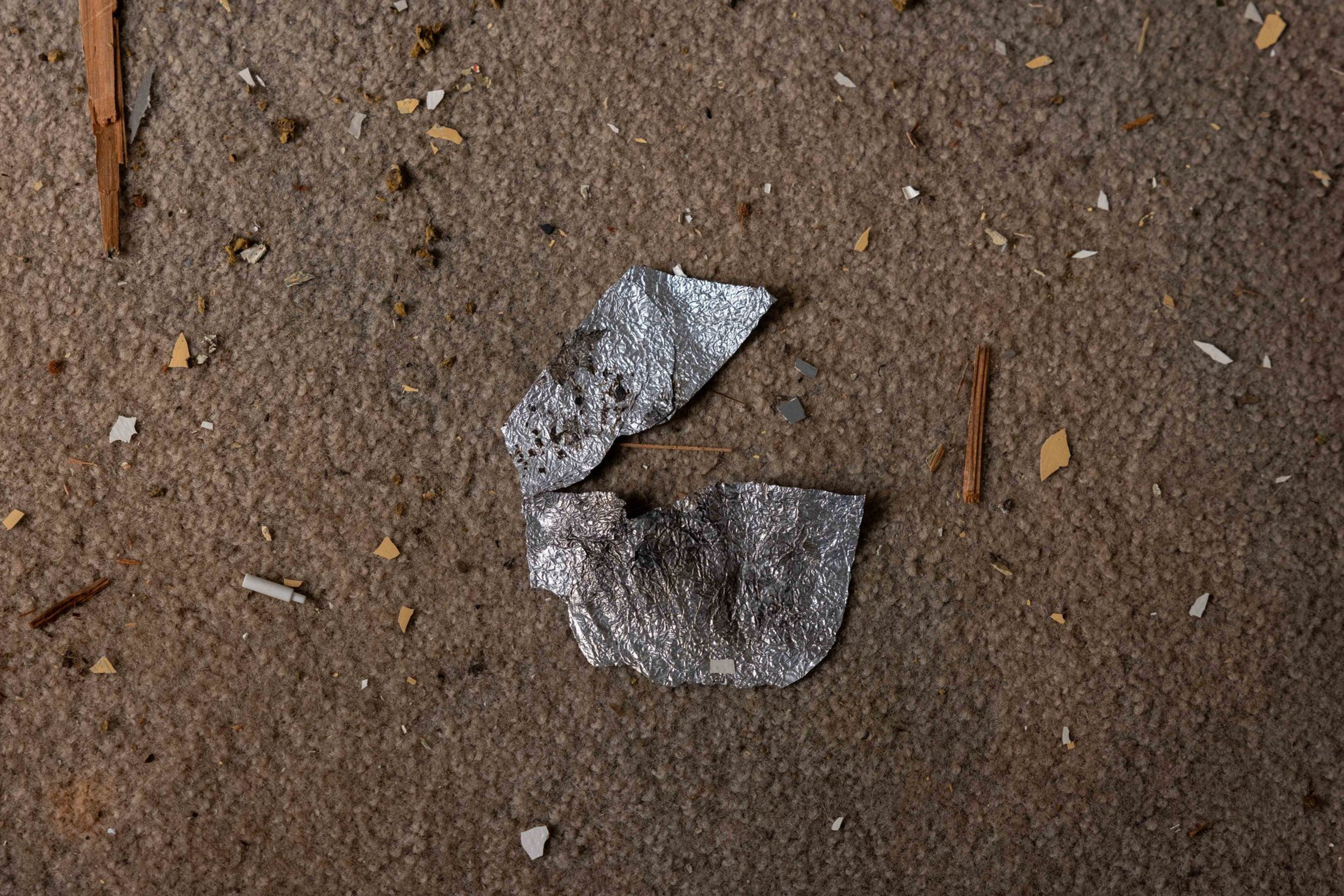 A torn, crumpled piece of silver foil lies on a carpet scattered with small debris like wood chips and tiny paper fragments.